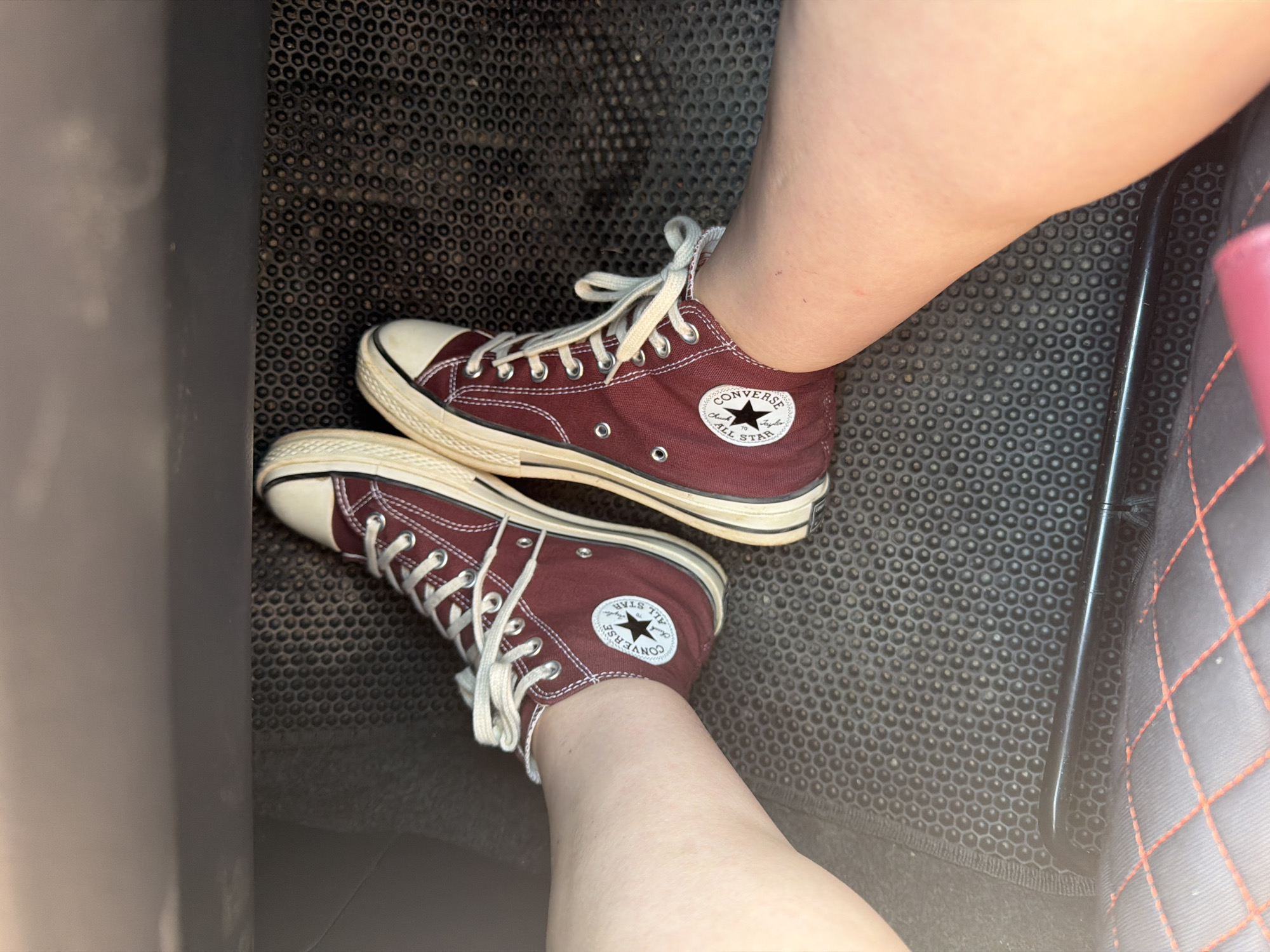 A top-down, close-up shot shows a pair of burgundy Converse Chuck Taylor All-Stars with white laces and soles on a person's feet, resting on a black, hexagonal-patterned floor mat inside a vehicle. The mat has a textured surface. The shoes are in focus, with the right shoe positioned slightly in front of the left. The surrounding environment includes dark gray interior car panels and a portion of a car seat with red stitching. The scene appears to be indoors, likely within a car. There is no information in the image to determine its location in the city of Chaikovsky, Russia.