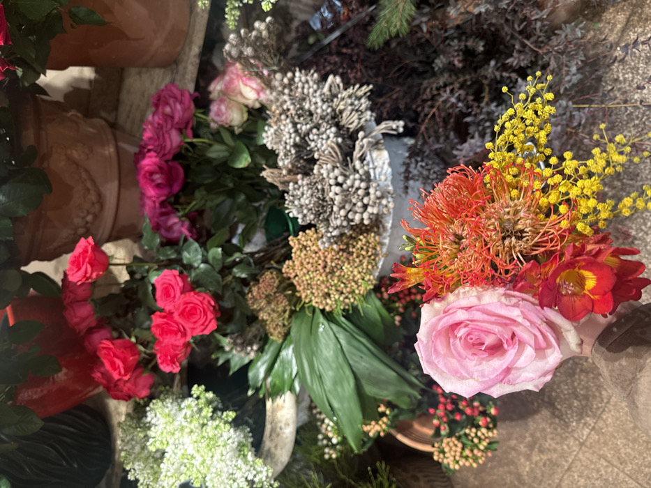 A vibrant collection of flowers in varying colors and textures is displayed. Pink roses, red freesia, and spiky orange pincushion protea mingle with feathery yellow mimosa. Other greenery and filler include silvery dried flowers and small white blooms. The arrangement sits amidst terracotta pots and a mix of other plant materials, creating a rich and diverse floral display.