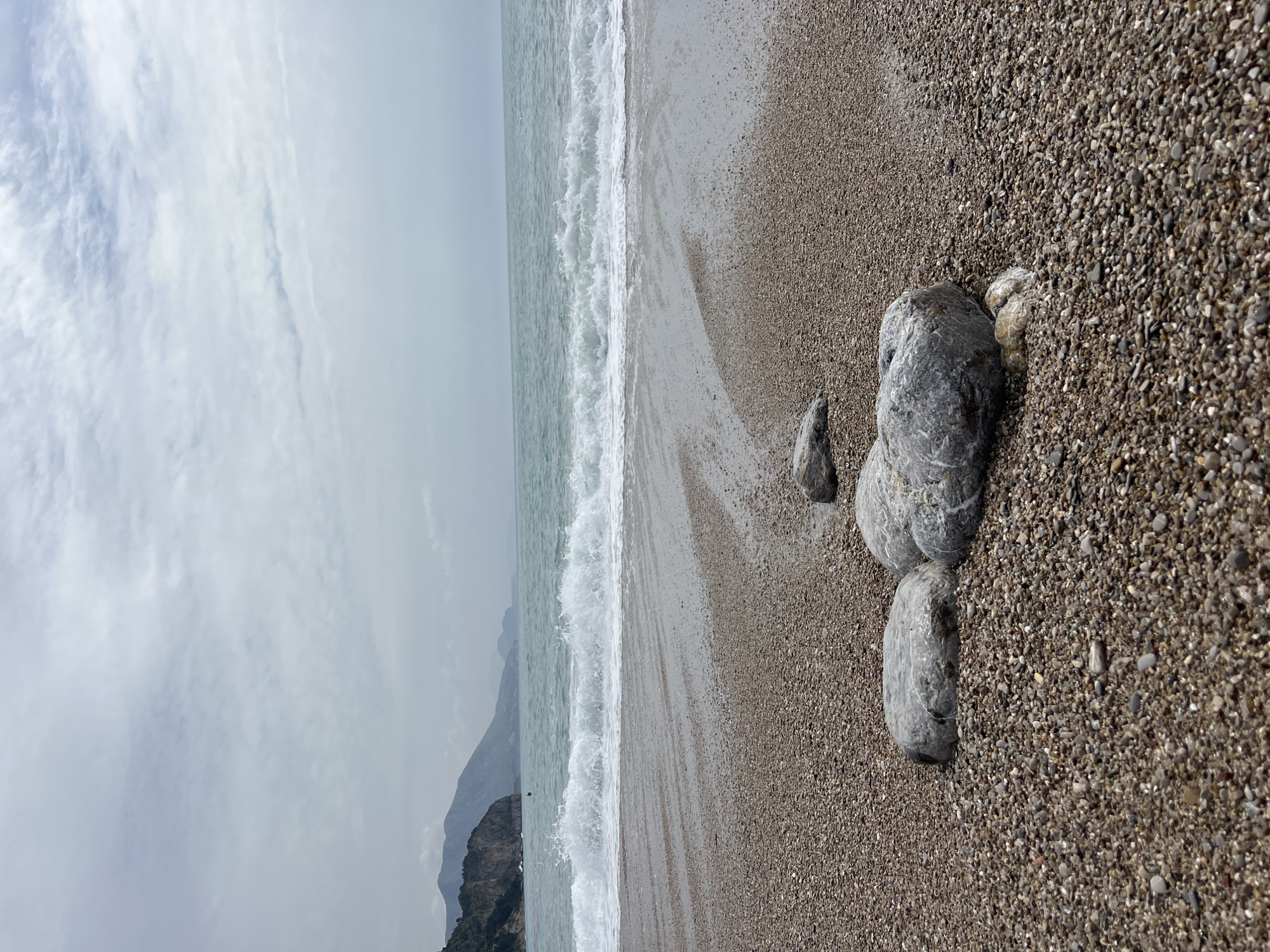 The image depicts a pebble beach on a coastline, likely near Prijevor, Montenegro, given the prompt. The sky is overcast. The sea has gentle waves breaking along the shore. In the foreground, several gray rocks are positioned on the pebble beach. A mountain is visible in the distance, partially obscured by clouds. The scene is static, with no visible people or actions taking place.