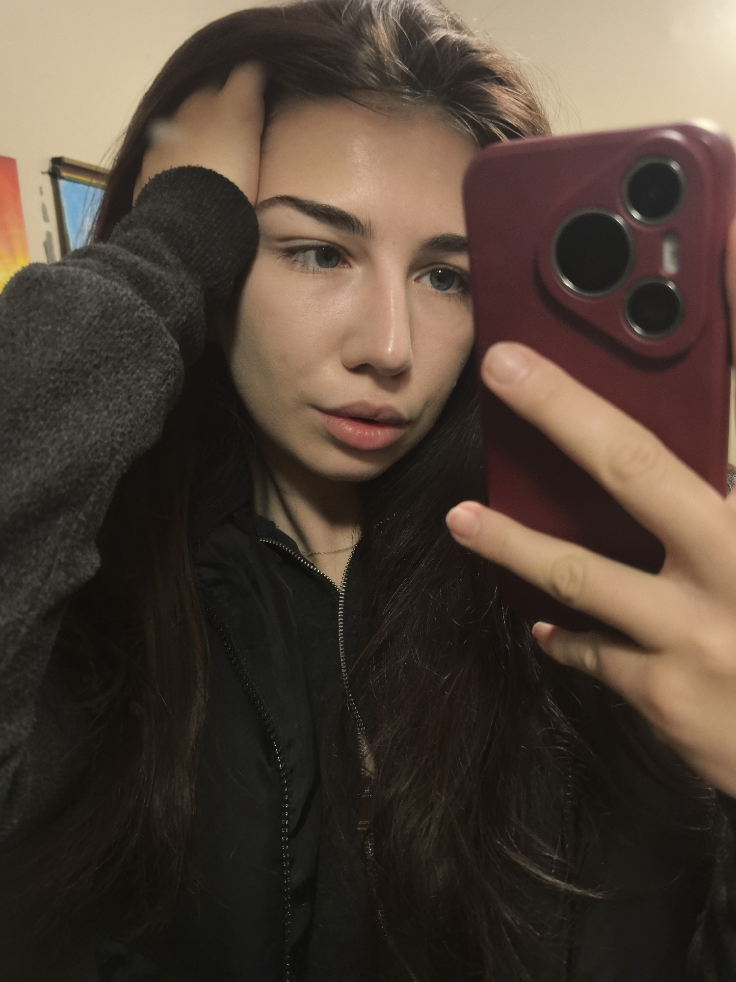 A young woman with long, dark hair and fair skin is taking a selfie with a red smartphone. She is wearing a gray hooded sweatshirt with a black jacket underneath. Her right hand is touching her forehead, and her left hand is holding the phone. The background is blurred, but a painting can be seen on the wall. The location in the city of Stara Zagora, Bulgaria is not discernible from the image.