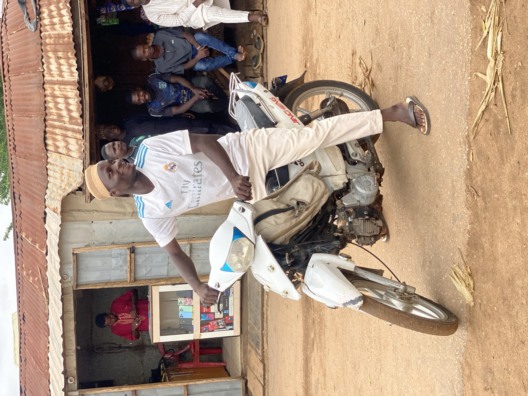A group of Nigerian men are gathered in front of a roadside shop with a corrugated metal roof in Rigachikun, Nigeria. The primary subject is a man in the foreground, seated on a white motorcycle, wearing a white Adidas jersey with "Fly Emirates" text and light-colored pants. He is smiling and looking towards the right. To his left, behind him, a man in a red garment is visible inside the shop, interacting with items on a display shelf. Several other men are seated or standing behind the motorcycle, in the background, some smiling. The shop appears to be a small electronics store, with cell phones displayed in a glass case. The ground is dusty and appears to be a dirt road.