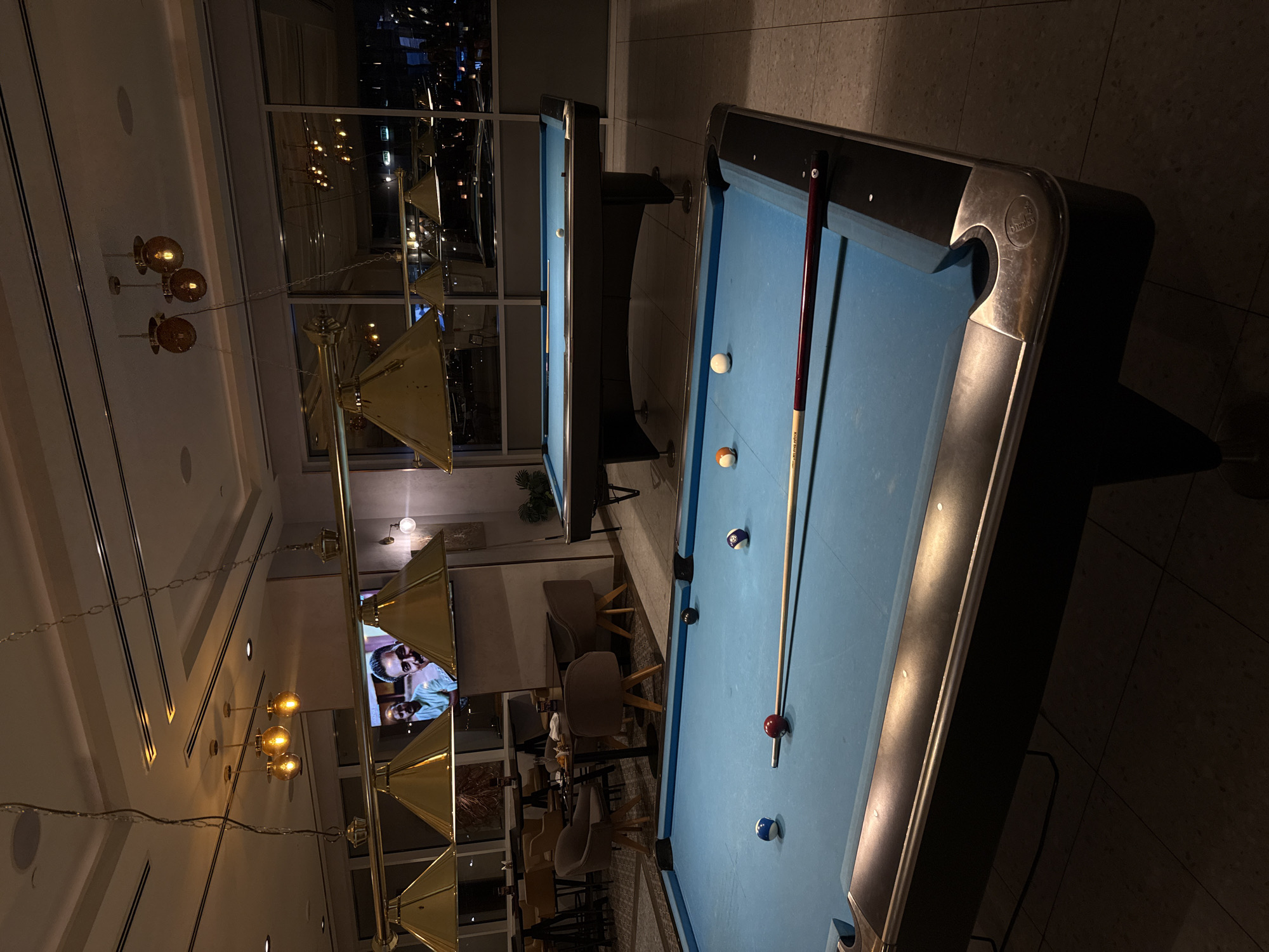 The image shows two pool tables in a room with a low-angle view of the Burj Khalifa through a window. The room has modern lighting fixtures and furniture, suggesting a contemporary interior design, but it is not known to be located in Dubai. Pool balls are positioned on one of the tables, accompanied by a cue stick. The setting implies a leisure or recreational area, likely inside a hotel or entertainment venue.