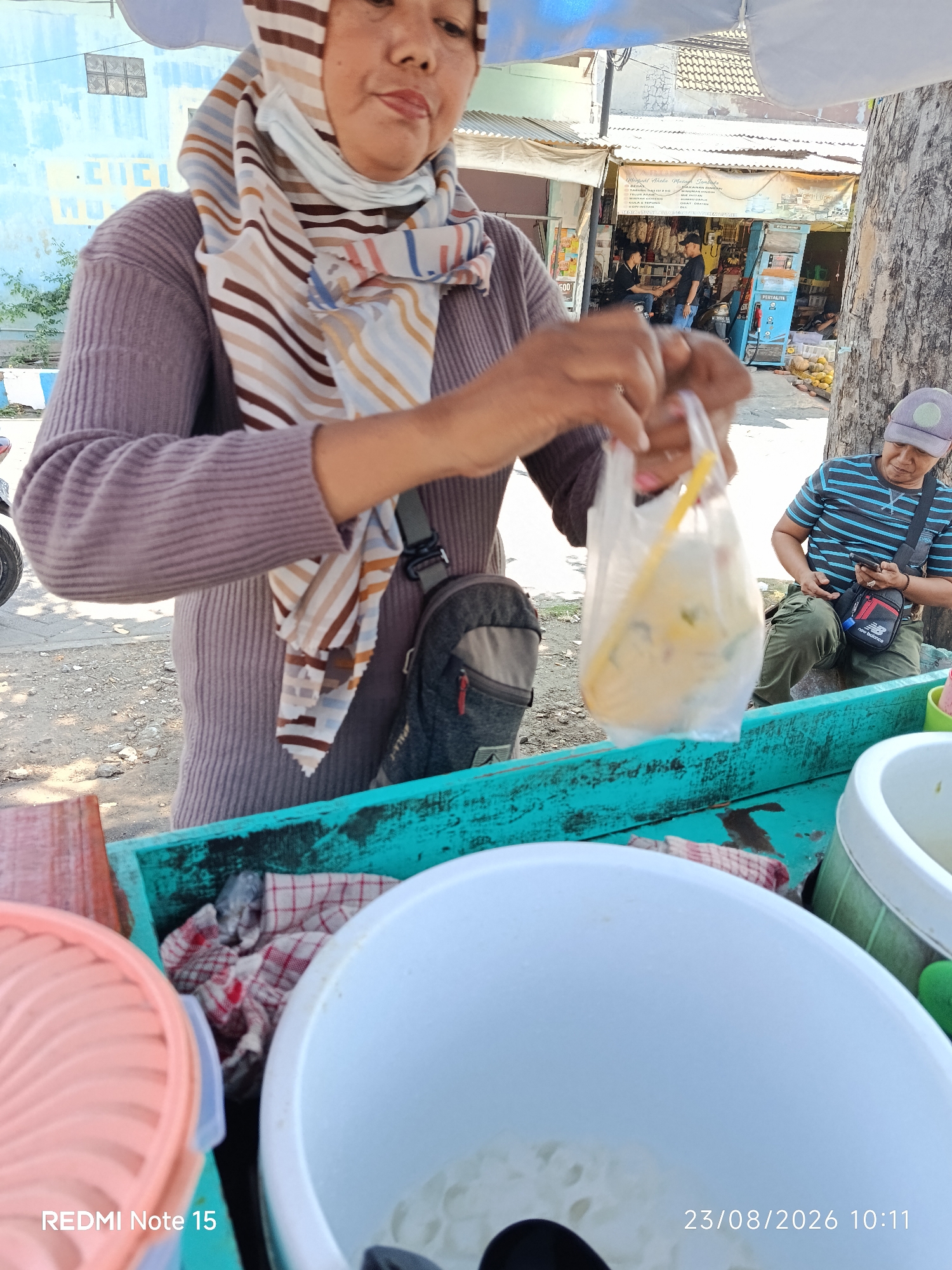 In Keboansikep, Indonesia, a woman wearing a purple long-sleeved shirt and a colorful striped hijab is standing behind a turquoise-colored wooden cart. She is holding a transparent plastic bag containing food, and her hands are engaged in preparing or handing over the item. A dark-colored sling bag is attached to her shoulder. To her right, a person wearing a striped blue and black shirt and a cap is seated, looking at their phone. The background reveals a street scene with buildings, a tree, and a shop displaying various goods. The cart itself contains a pink food container on the left and a large white bowl filled with white food items on the right. A red and white checkered cloth is placed on the cart. The timestamp "23/08/2026 10:11" and the text "REDMI Note 15" are visible in the lower part of the image.