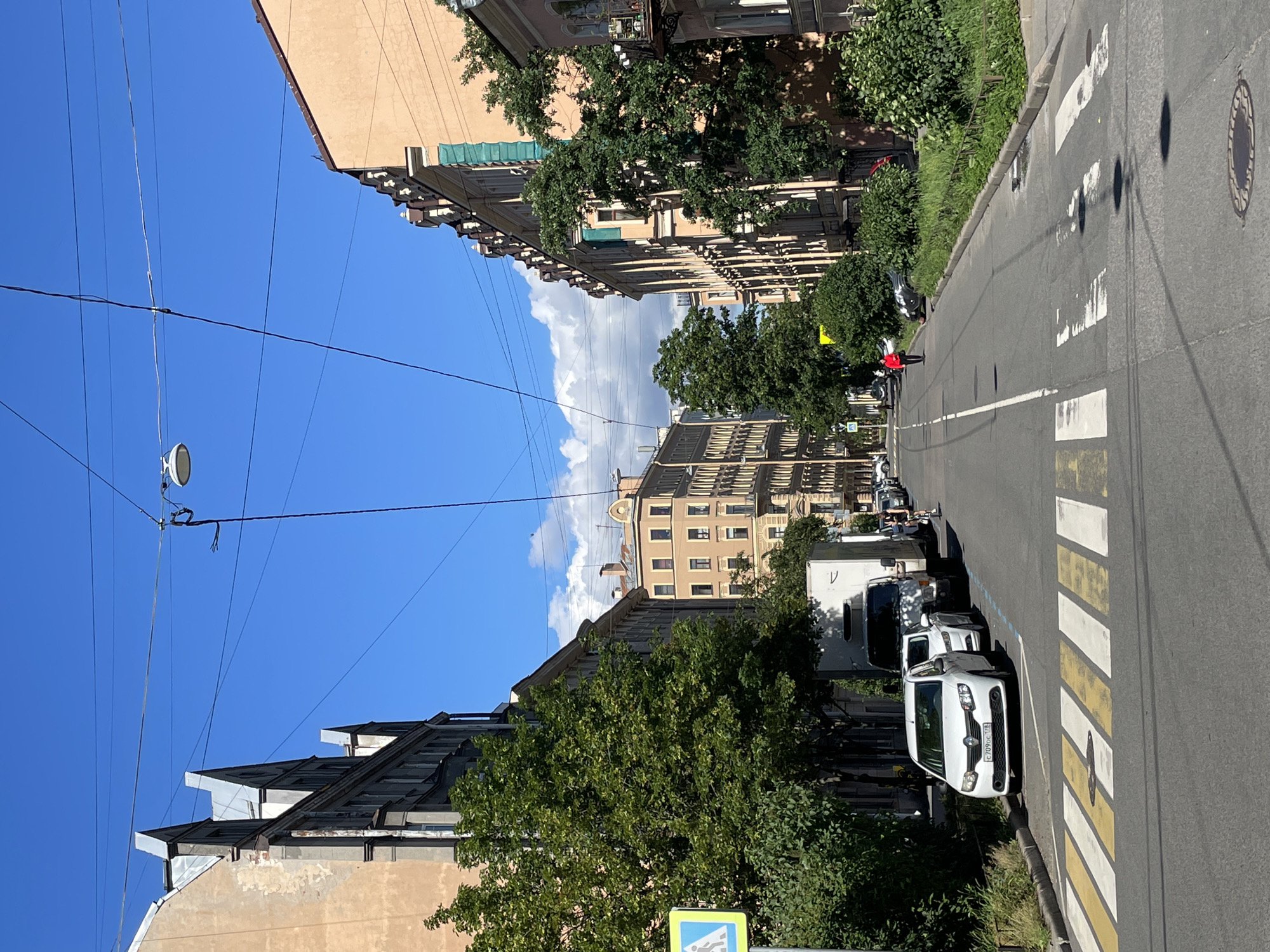 This is a vertical, eye-level, outdoor shot of a street scene in Saint Petersburg, Russia. The image is dominated by buildings on either side of a road, with a bright blue sky and scattered white clouds above. The architecture appears to be historic, with ornate details on the building facades. Lush green trees line the sidewalk, casting shadows on the road.
The road itself is paved and has a pedestrian crossing marked with white and yellow stripes. Several vehicles are parked along the side of the road, including a white van and a few other cars. A person in a bright red jacket is walking across the pedestrian crossing. The lighting suggests it is daytime, likely mid-morning or afternoon, with bright sunlight. The overall impression is a clear, sunny day in an urban environment with a mix of old-world charm and modern life.
There is some visible text on the license plate of the white car parked nearest the pedestrian crossing. It reads "C 789PC 198". There is also a blue and yellow traffic sign visible in the lower left corner of the image, though the text on it is illegible.