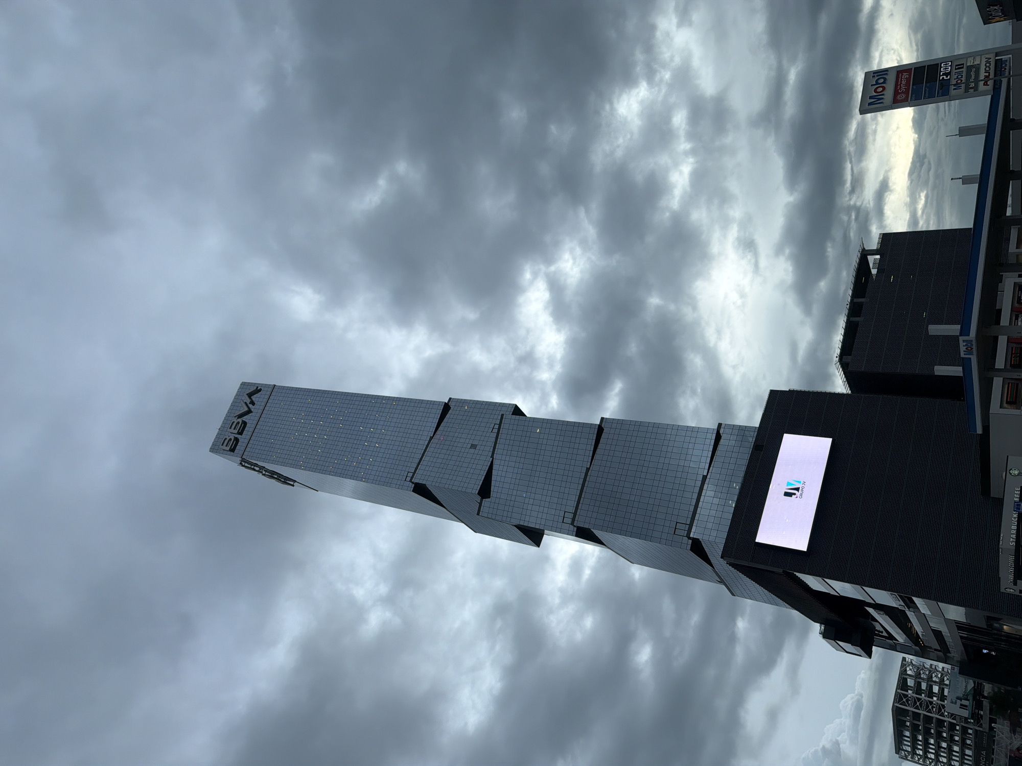 The image shows a modern, angular skyscraper with the "BBVA" logo prominently displayed at its peak. The skyscraper is clad in glass panels and appears to be under a cloudy sky. In the foreground, there is a dark, multi-story building with a large illuminated screen displaying the "JV GRUPO JV" logo. To the right, a Mobil gas station sign with prices is visible. The overall scene suggests an urban environment with contemporary architecture. This location is in San Bernardino Tlaxcalancingo, Mexico.