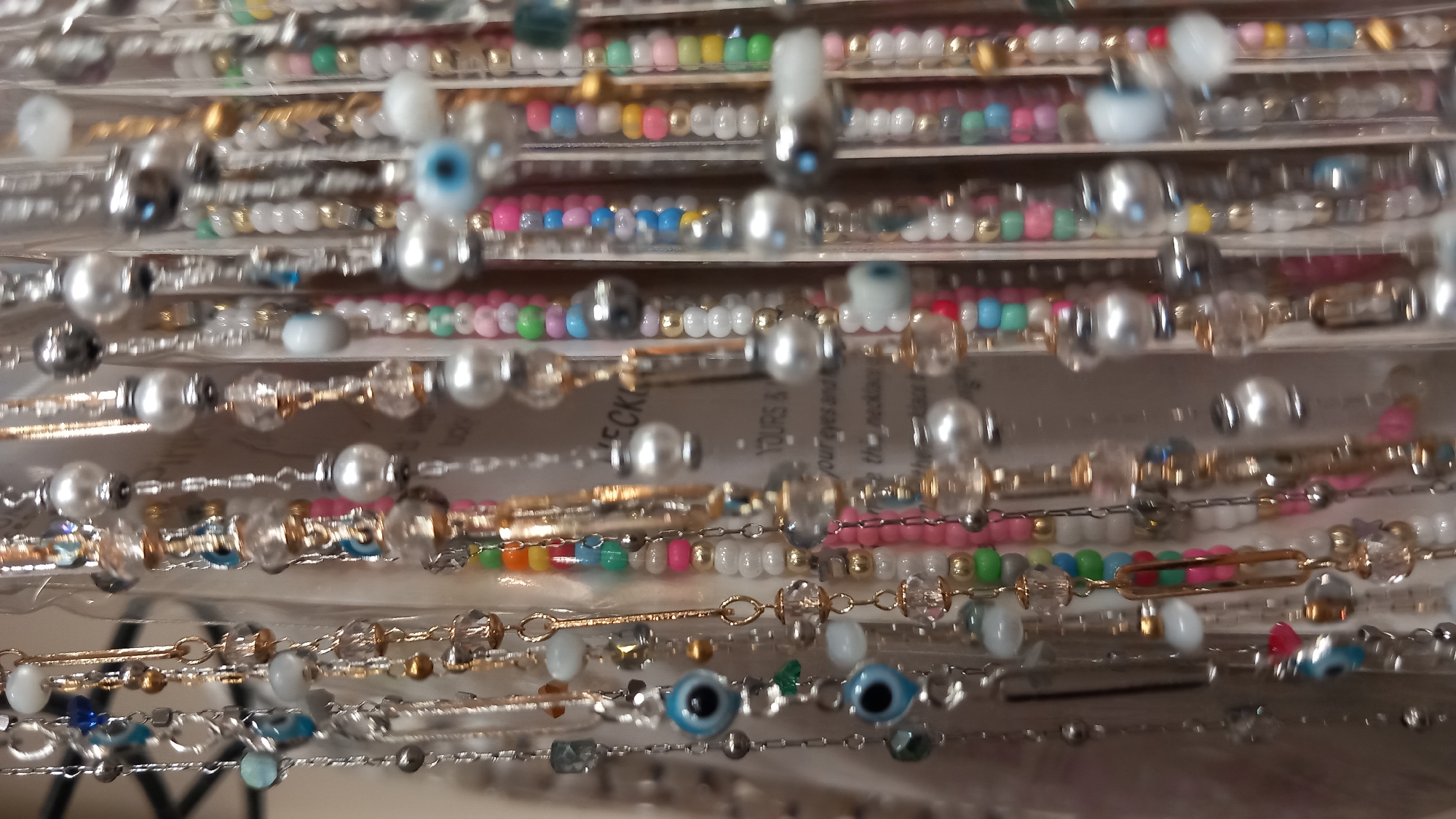 A close-up, shallow depth-of-field photograph shows multiple strands of colorful beaded jewelry, possibly necklaces or bracelets, hanging vertically. The jewelry consists of a variety of beads including pearls, opaque colored beads in pink, green, blue, and yellow, and clear or iridescent crystal beads. Some strands also feature metallic beads in silver and gold tones. Several strands include charms, most notably blue and black evil eye charms. The background is out of focus and appears to be shelving or a display, with some text visible, potentially a product label or sign. The lighting is bright and diffused. There are no people visible in the image. The scene is not identifiable as being in the city of Kos, Greece, or any specific location.