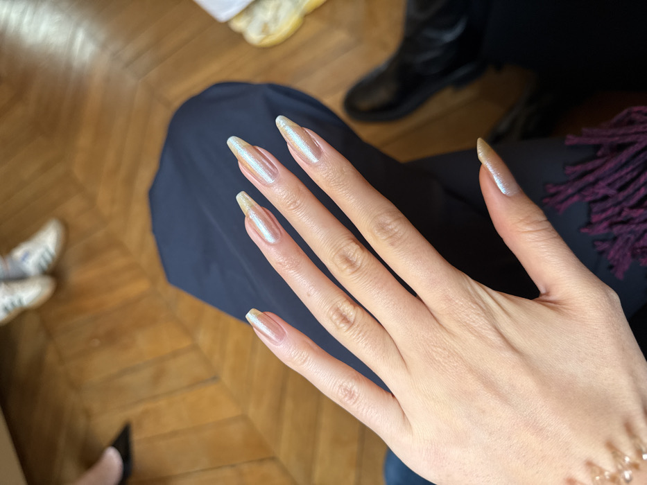 The image displays a close-up, downward-angled view of a person's left hand prominently featuring manicured nails. The nails are long, with an almond-coffin shape, and exhibit an iridescent, pearlescent finish that shifts between a light pinkish-gold base and a pale blue-green shimmer. The hand rests on dark blue fabric, likely clothing worn by the subject, with a glimpse of dark red or purple fringed textile visible towards the bottom left.
The background, largely out of focus, reveals a polished light-brown wooden parquet floor laid in a classic herringbone pattern. This type of flooring is characteristic of older, elegant interiors commonly found in apartments and buildings within Paris, France. Various partial views of footwear belonging to other individuals are visible in the blurred background, suggesting multiple occupants in the room. These include a black pointed shoe (top left), white athletic sneakers with three stripes (top center), and a black formal leather shoe (bottom right). A yellowish-white blurred object is also visible near the formal black shoe. The scene depicts an indoor setting.