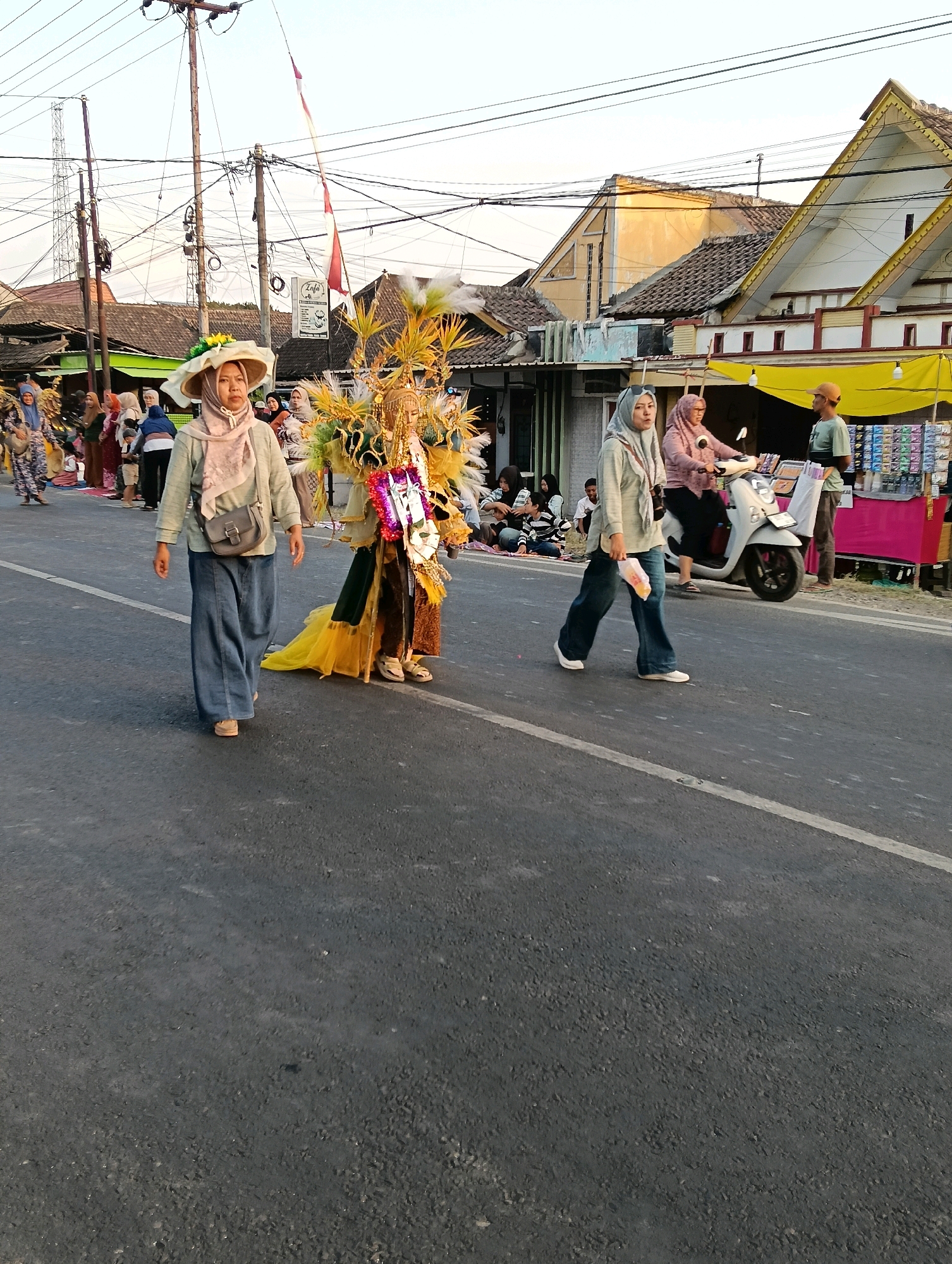 In Kasiyan, Indonesia, a street scene unfolds with a procession or parade. In the foreground, a person in a large, elaborate costume, adorned with gold and green, walks down the center of the asphalt road. To the left, a woman wearing a wide-brimmed straw hat, a light-colored top, and wide-leg jeans walks alongside the costumed figure, carrying a gray crossbody bag. To the right, another woman, also in a hijab and light-colored top with jeans, walks further down the road. Behind them, a white scooter is being ridden by a person in a pink hijab, and a man in a light green shirt and a baseball cap stands near a stall with various items displayed. The background shows buildings with tiled roofs and utility poles with numerous wires crisscrossing overhead. The sky is overcast and bright. Several other people are visible in the background, some seated on the side of the road, observing the event.