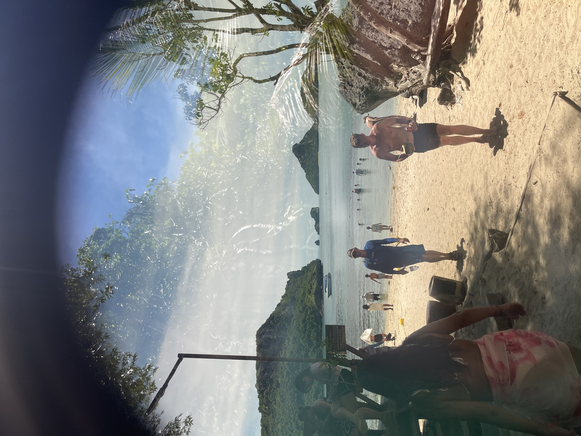 The image depicts a tropical beach scene on a sunny day. In the foreground, people are walking on the sand, some are wading in the shallow water, and others are seated on a wooden structure. A woman in a bikini with a sarong tied around her waist walks away from the camera, while a shirtless man in black shorts walks towards the camera holding a coconut and a can. In the background, several islands covered in lush green vegetation rise from the clear blue water. A white boat is visible near one of the islands. The sky is a bright blue with some scattered clouds. The image appears to have a double exposure or a reflective surface overlay, as there are translucent images of trees and foliage superimposed over the beach scene, particularly in the upper half of the frame. The overall impression is one of a busy tourist destination in Thailand, likely an island within a national marine park.