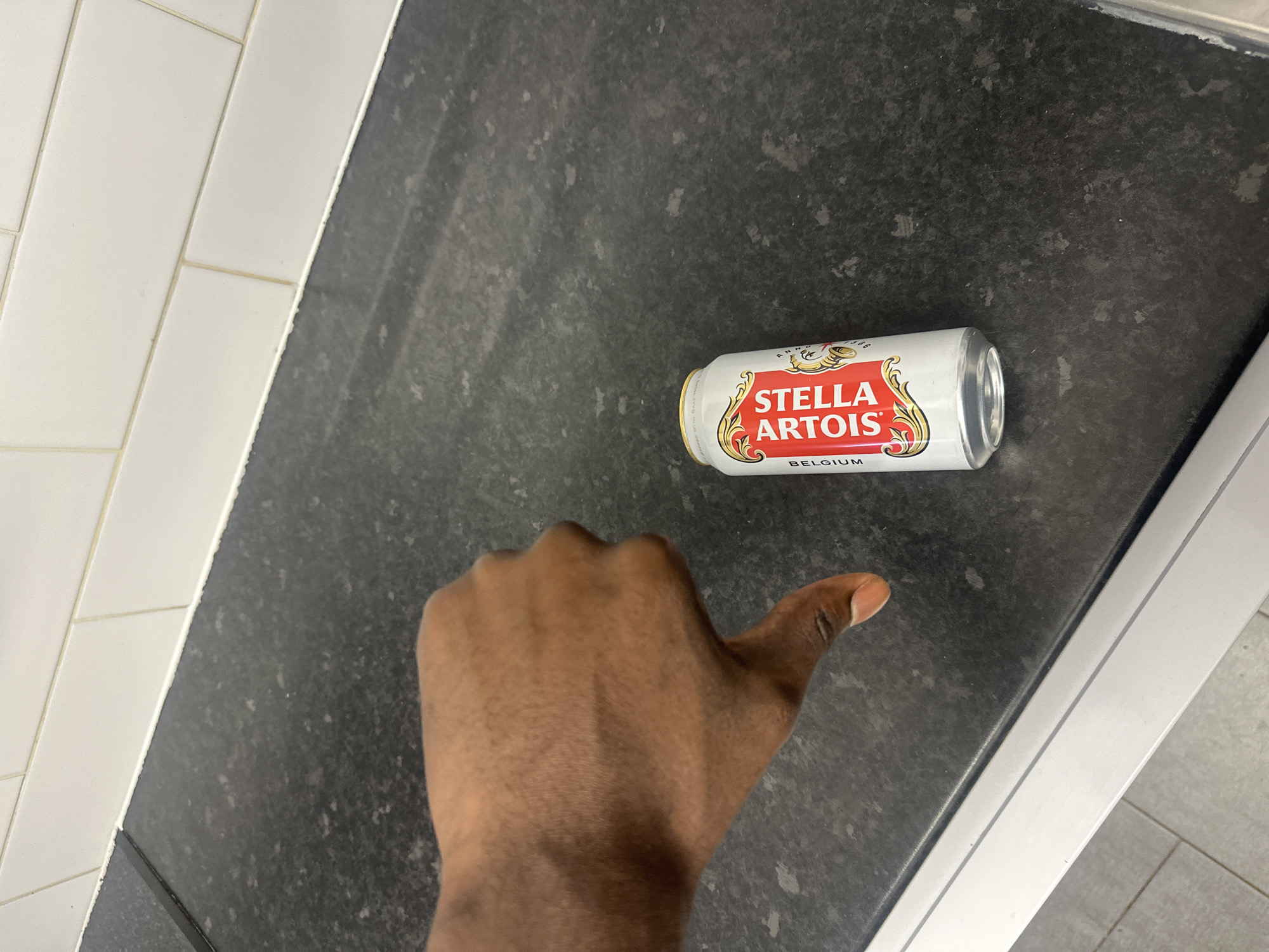 A silver can of Stella Artois beer lies on a dark grey countertop. A dark-skinned hand gives a thumbs up in front of the can. The countertop is next to white tiles. The location is a kitchen or bathroom with tile and a countertop. No information to identify Canterbury.