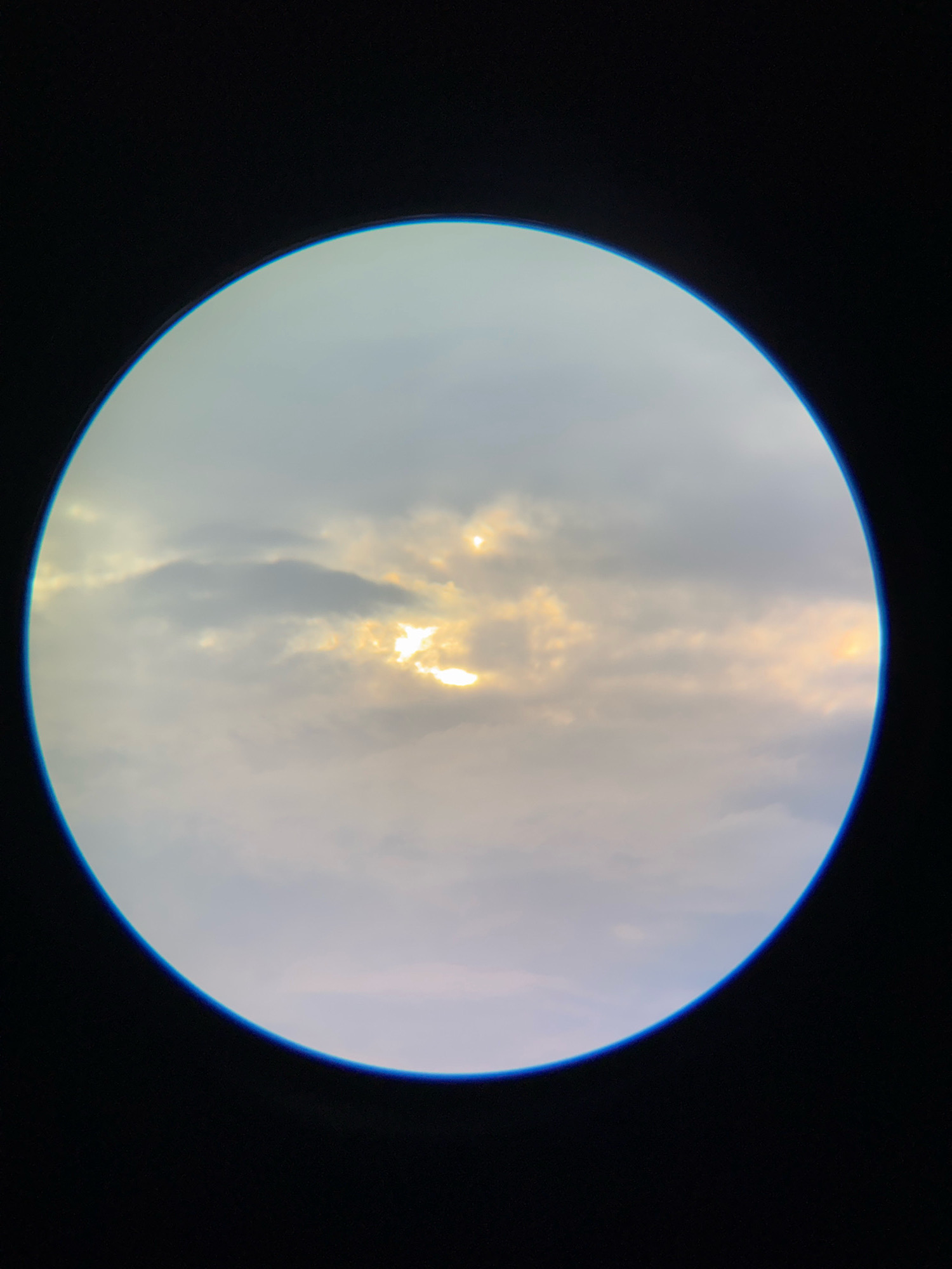 A photograph taken through a circular lens shows a cloudy sky with the sun partially visible. The sky is a mix of light gray and pale yellow hues, with some darker clouds obscuring patches of the sun. The sun's rays are breaking through in a few areas, creating bright spots of light. The edges of the circular frame are dark, with a faint blue glow around the inner rim. The image does not depict any specific location within Nha Trang, Vietnam, or any people or objects.