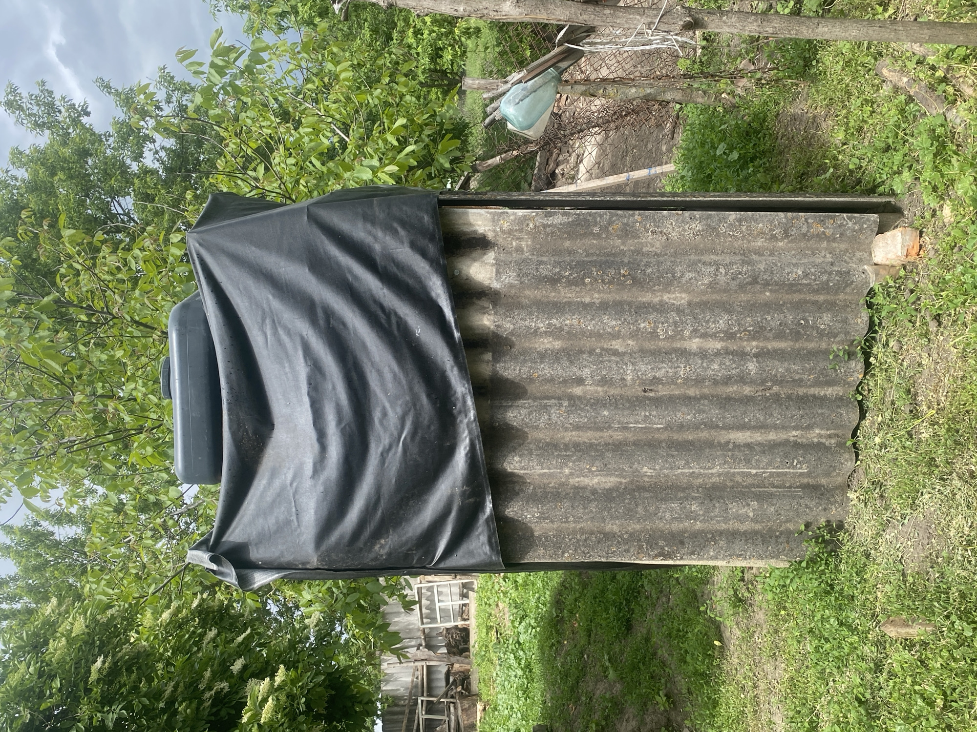 The image shows a corrugated asbestos-cement roof partially covered by a black tarpaulin, possibly protecting a water tank from rainwater. The surrounding area consists of foliage, trees, grass, and a fence, indicating a rural or semi-rural environment. A broken wooden structure can be seen in the background. The location is not discernable from the image.