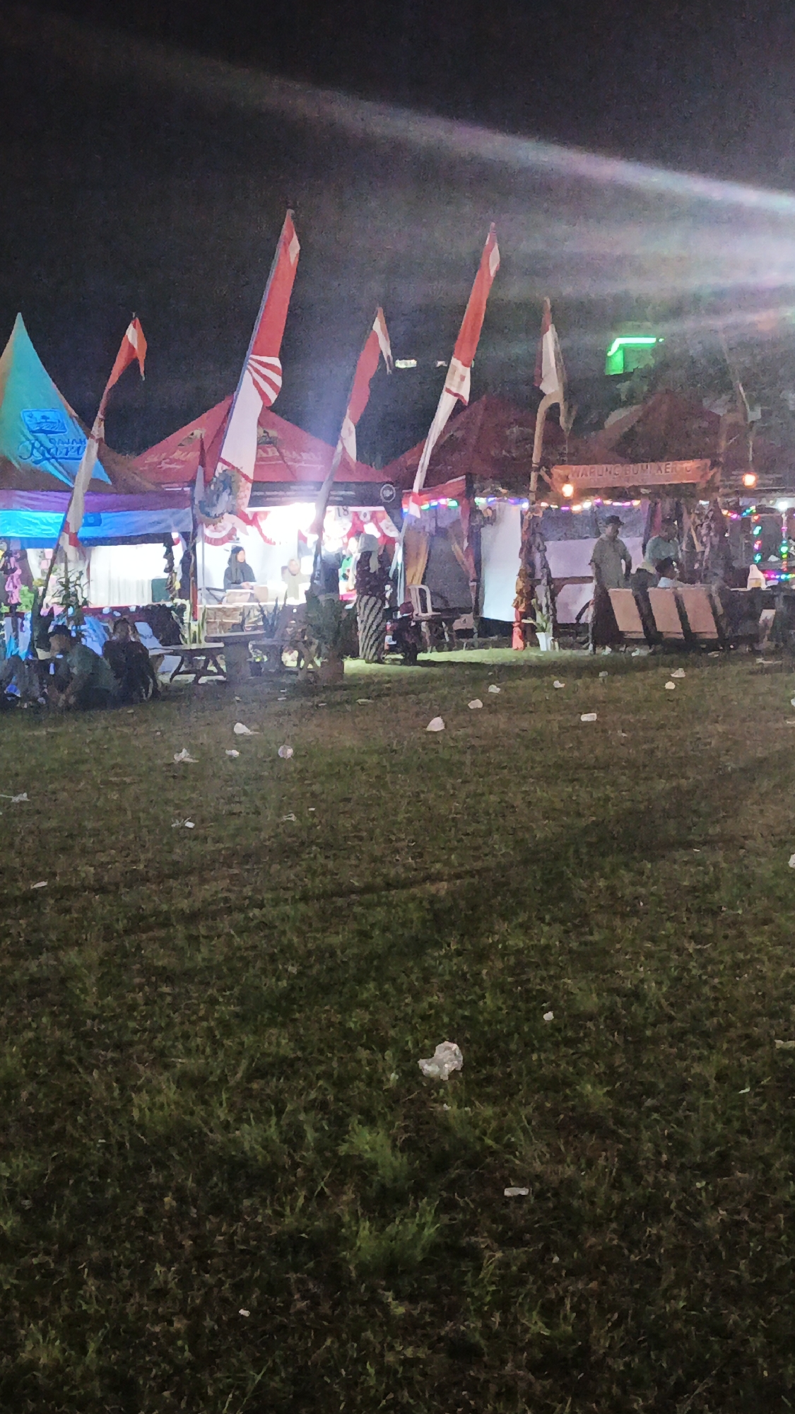 The image depicts an outdoor scene at night, likely a market or festival in Ketemasdungus, Indonesia, with a focus on several stalls and a grassy area in the foreground. In the background, there are multiple tents or stalls set up, adorned with Indonesian flags. People are visible at the stalls, some standing and interacting, while others appear to be seated. The lighting is artificial, with some bright lights illuminating the stalls and creating reflections in the sky. The foreground consists of a grassy field with scattered litter, suggesting a public gathering. A few people are seated on the grass in the lower left of the image. The overall atmosphere appears to be festive or communal.