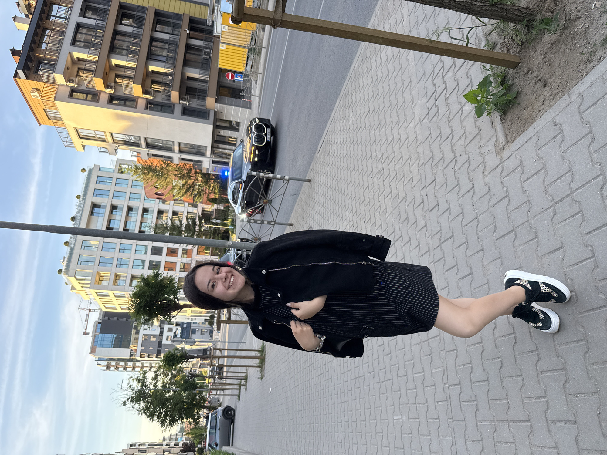 A woman is standing on a grey brick sidewalk in Sofia, Bulgaria. She is smiling, wearing a black jacket, a striped dress, and black and white sneakers. Behind her is a street with modern buildings. A black car with blue lights is parked on the road. Trees line the sidewalk, and the sky is partly cloudy.