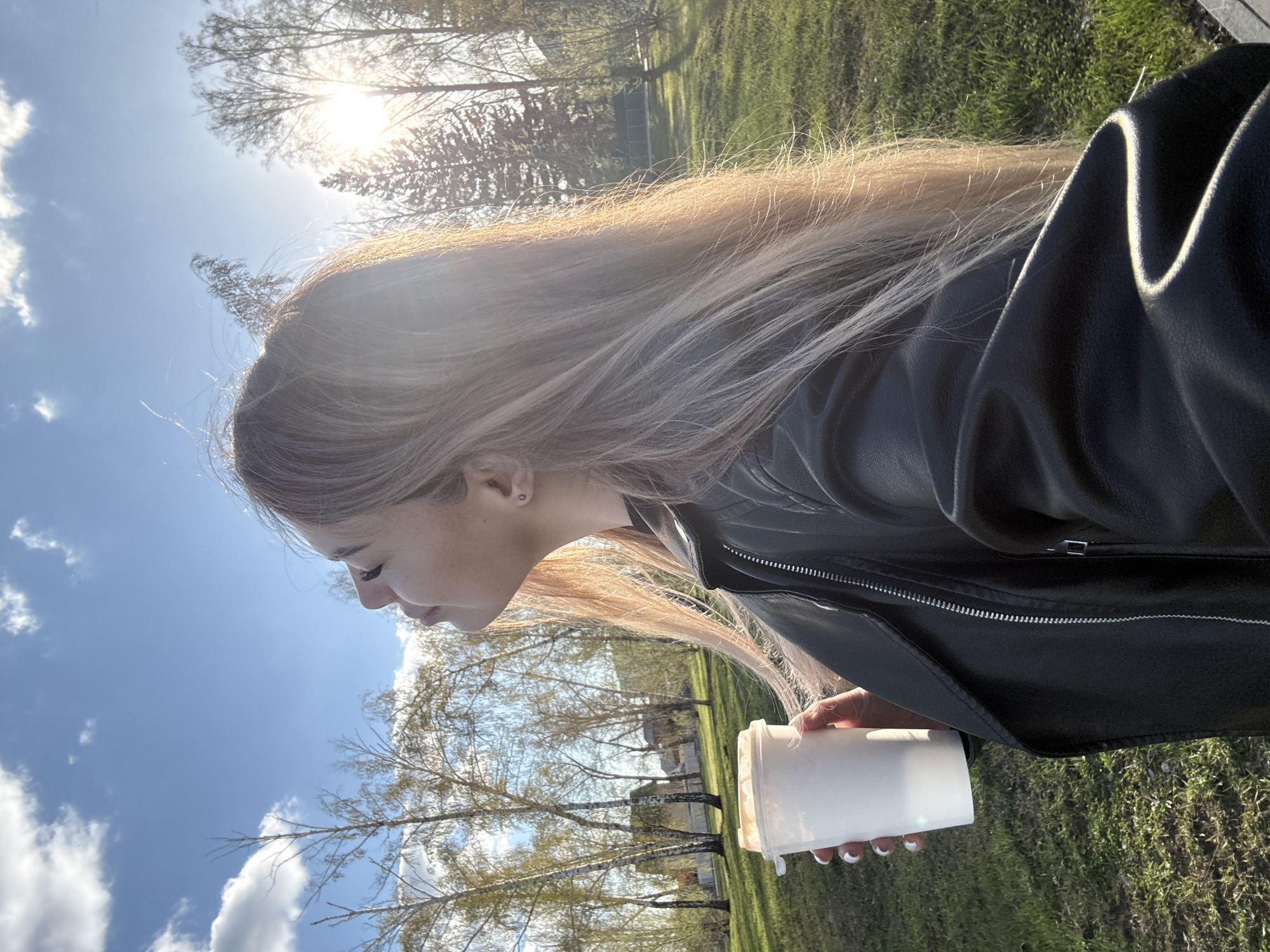 The image captures a profile view of a person outdoors on a sunny day. The subject is a young woman with long, light-colored hair, which appears blonde or light brown, illuminated by strong sunlight from above and behind her. She is wearing a black leather jacket. Her gaze is directed slightly downwards. In her right hand, she holds a plain white, opaque cup or bottle.
The setting appears to be a park or an open green space, characterized by green grass in the foreground and background, and several bare or sparsely leafed trees, suggesting early spring or late autumn. The sky is bright blue with scattered white clouds. The sun is prominently visible in the upper part of the frame, casting a strong glare and creating bright highlights, particularly on the subject's hair and jacket. The overall atmosphere is bright and clear.
No specific activity beyond holding the cup and looking is discernible. There is no visible text in the image.