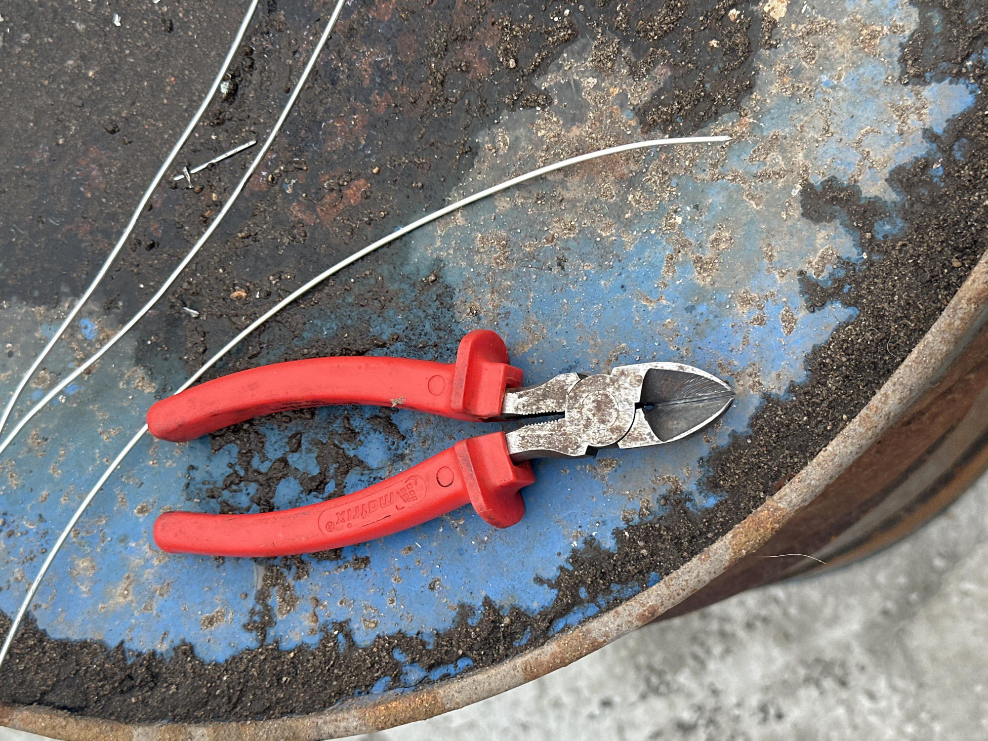 A pair of red-handled wire cutters rests on a blue, weathered surface of a barrel. The barrel's surface is stained with dark, earthy material and scattered debris, with streaks of rust visible along the edge. Several thin, metallic wires are laid across the surface, some appearing cut. The background is out of focus, suggesting an outdoor environment. There are no people or animals visible in the image. The scene appears to be an industrial or work area. The location is in the city of Tobyl, Kazakhstan.