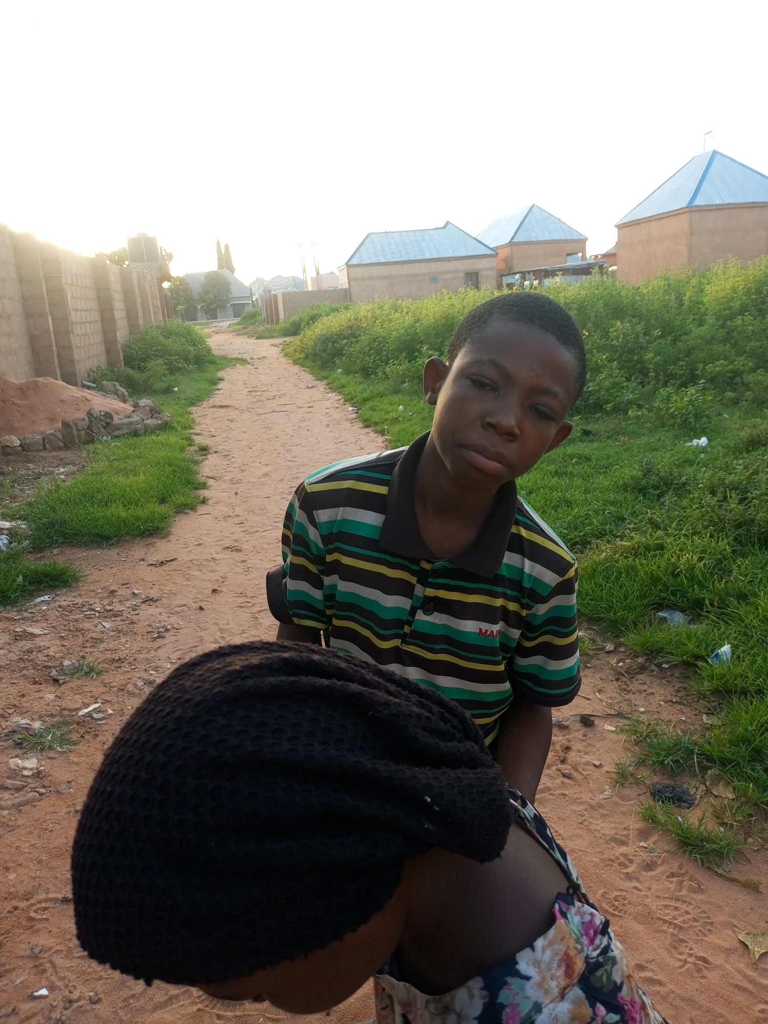 This media file depicts a medium shot of two people outdoors in what appears to be a rural or semi-rural area in Tempure, Nigeria. The subject in the foreground is a child, seen from the upper chest up, with a black knitted cap covering their head. The child's face is partially visible, turned towards the camera with a neutral expression. They are wearing a horizontally striped short-sleeved shirt with colors of green, yellow, black, and white.
Behind the child, another individual is partially visible, their upper body facing away from the camera. This person is wearing a dark garment, possibly a shirt or jacket, and their arm is visible on the right side of the frame.
The setting is a dirt path or road, with dry, sandy soil. To the left of the path is a low wall made of cinder blocks, with a pile of sand and some rocks next to it. Beyond the wall, there are some green bushes and trees. To the right of the path, more lush green vegetation is visible. In the background, several buildings with light brown walls and distinctive blue-tiled roofs are present. The sky is bright, suggesting it is either early morning or late afternoon, with the sun casting a warm glow. The overall atmosphere appears calm and ordinary. There is no discernible activity or event taking place, it seems to be a posed or candid photograph of individuals in their environment. No visible text is present in the image.