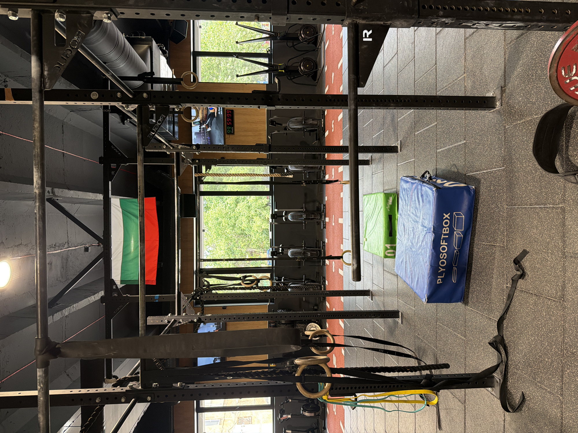The image depicts the interior of a gym. The scene is dominated by a large, black metal squat rack structure, which occupies a significant portion of the frame. Various gym equipment is attached to or positioned around the rack, including gymnastic rings, ropes, and a blue plyometric box with the text "PLYOSOFTBOX" printed on its side. To the left of the squat rack, a Bulgarian flag hangs, its white, green, and red stripes clearly visible.
In the background, large windows offer a view of lush green trees, suggesting an outdoor environment beyond the gym walls. Through the windows, rowing machines are also visible, indicating the diverse range of equipment available in the facility. The floor is covered with dark grey textured tiles. The lighting appears to be artificial, with a bright circular light fixture visible on the left side of the image. There are no people visible in the image.
The overall impression is that of a well-equipped fitness center, possibly a CrossFit gym, given the presence of a squat rack and plyometric boxes. The focus is on the gym equipment and the structural elements of the facility, with the Bulgarian flag adding a specific national or local context. The scene is static, with no discernible activity taking place.