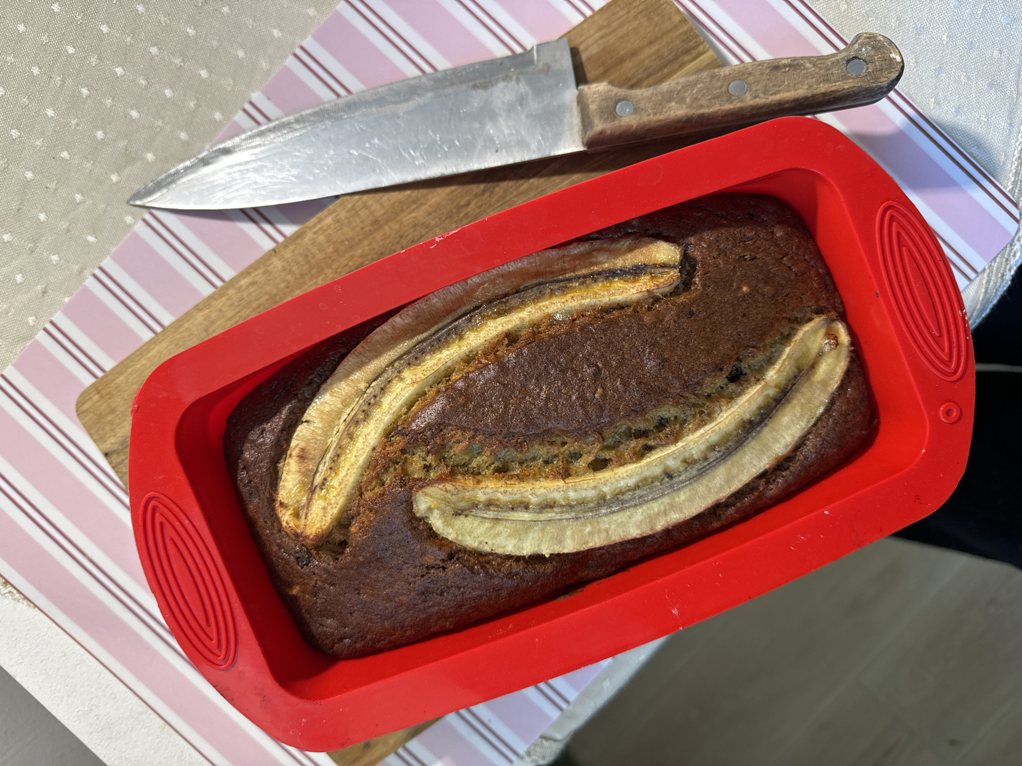 The image shows a freshly baked banana bread in a red silicone loaf pan, sitting on a wooden cutting board. A large knife with a wooden handle rests on the cutting board next to the banana bread. The cutting board is placed on a surface with a striped pink and white pattern. The banana bread has two banana halves placed on top as decoration. The environment appears to be a kitchen or dining area. There are no people visible in the image. There is no evidence to connect this image to Krasnoyarsk, Russia.
