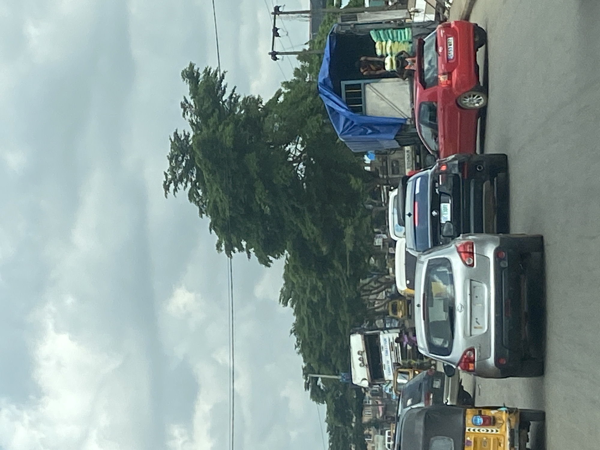 The image captures a daytime street scene in Egan, Nigeria, depicting a line of vehicles in what appears to be traffic. The perspective is from inside a vehicle, looking forward.
In the foreground, a silver hatchback is visible, followed by a black car and a yellow three-wheeled vehicle (possibly a tricycle taxi or "keke napep"). Further down the road, a red hatchback is stationary, followed by a truck with a blue tarp covering part of its load area. Inside the truck, a person is visible next to stacks of light-colored bags, possibly containing goods or produce. Beyond the immediate vehicles, more cars and trucks are visible in the distance, indicating a busy road.
The right side of the image features a line of lush green trees, suggesting a tree-lined street or area of vegetation. Utility poles with overhead wires run along the side of the road. The sky is overcast with gray clouds, implying a cloudy day.
Visible text includes license plates on several vehicles. The red hatchback displays "KBM 592 KY". A black car further back has a partially visible plate that appears to be "KSM 13DF". On a white truck in the mid-distance, faint text that might read "CONGO TRADES" is barely discernible. The overall atmosphere suggests a typical day on an urban or semi-urban road with moderate traffic.