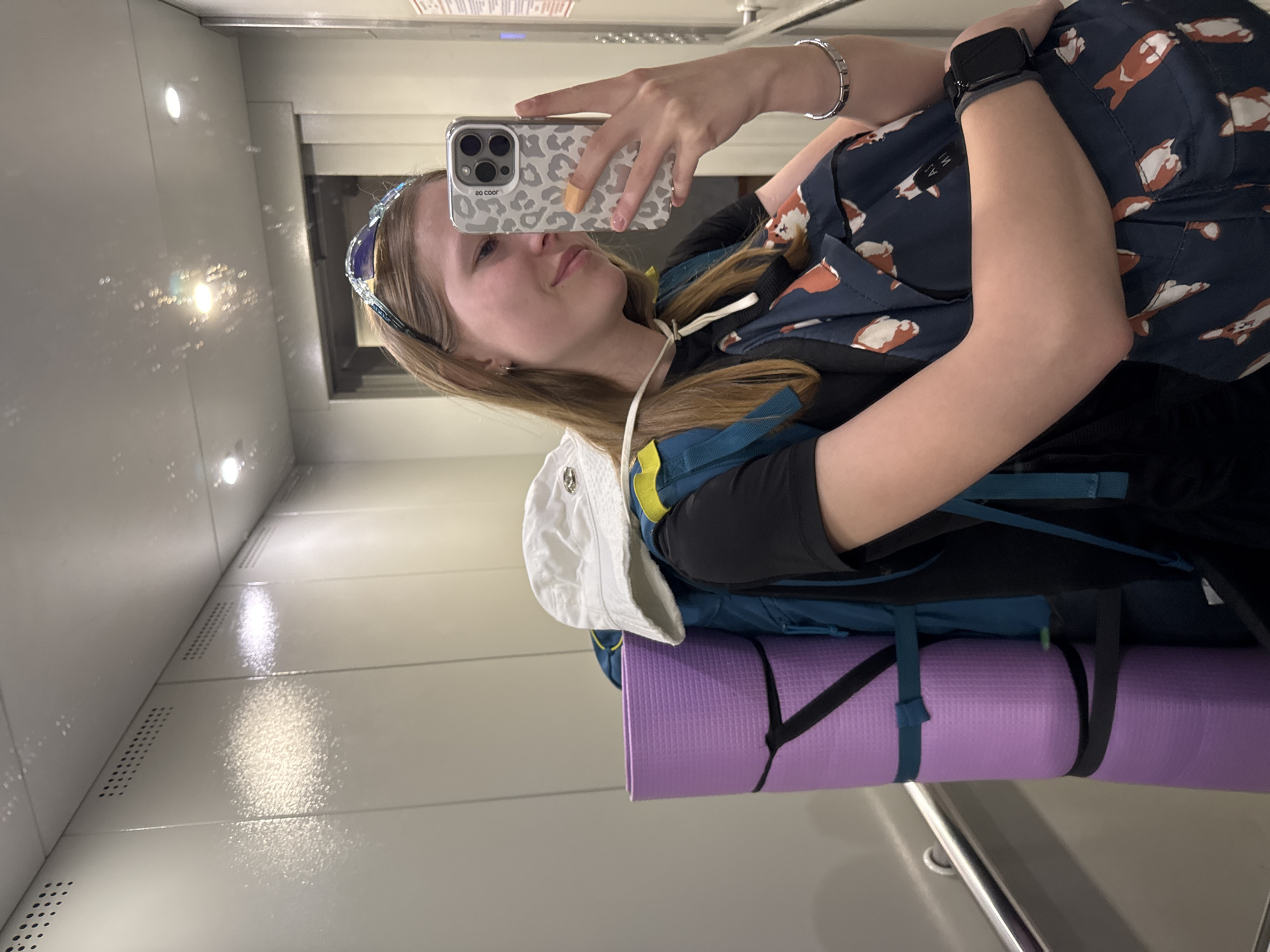 A young woman is taking a selfie in what appears to be an interior space, possibly a train car. She is wearing a black long-sleeved shirt, a headband, and has a backpack on. The backpack has a light-colored hat attached and a rolled purple yoga mat. She has an Apple Watch on her left wrist. Her phone case has a leopard print design. The environment appears to be clean and modern, with light gray walls and metallic accents. Based solely on visual details, there's no discernible information to confirm the location in Chaikovsky, Russia, or any details beyond the immediate scene.