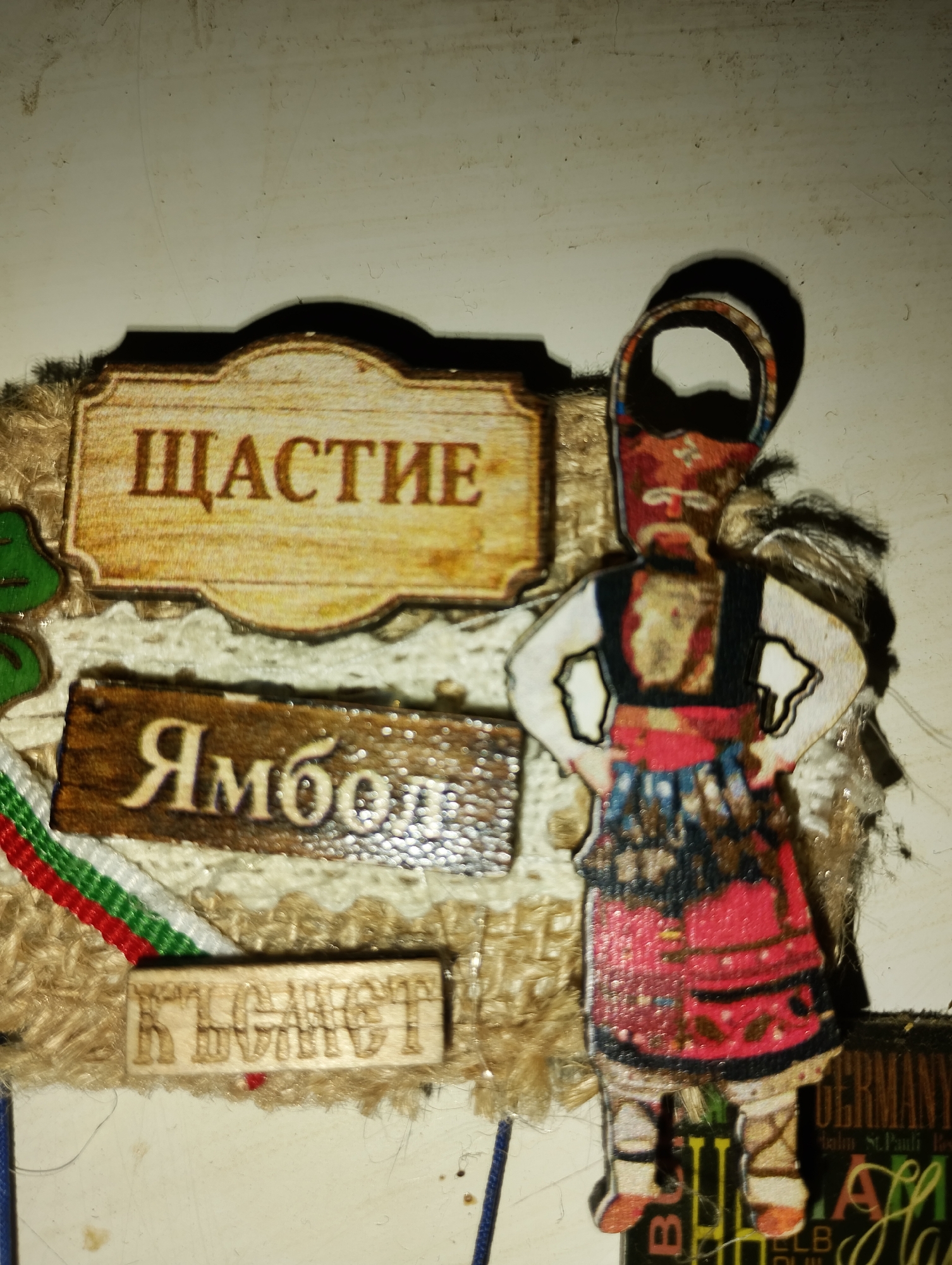 An overhead close-up shot shows a crafting project featuring a cutout of a traditionally dressed Bulgarian doll. The doll is positioned on the right, facing left with hands on hips. Behind the doll, on a light-colored surface, are two wooden signs with Cyrillic text. The top sign, a light tan wood with a scalloped edge, reads "ЩАСТИЕ" (meaning "happiness") in dark brown, embossed text. Below it, a darker, rectangular wooden sign reads "Ямбол" (a city in Bulgaria) in cream-colored, embossed text. To the left of the doll, a green leaf cutout is visible, as well as a strip of the Bulgarian flag colors (white, green, red). The bottom of the image shows a small, light-colored wooden rectangular sign with embossed Cyrillic text that is difficult to read clearly, but appears to be "бъдете" (meaning "be"). The doll is adorned with a red, blue, and black patterned skirt and a red sash, and wears a headpiece. The overall impression is of a decorative piece with a Bulgarian theme.