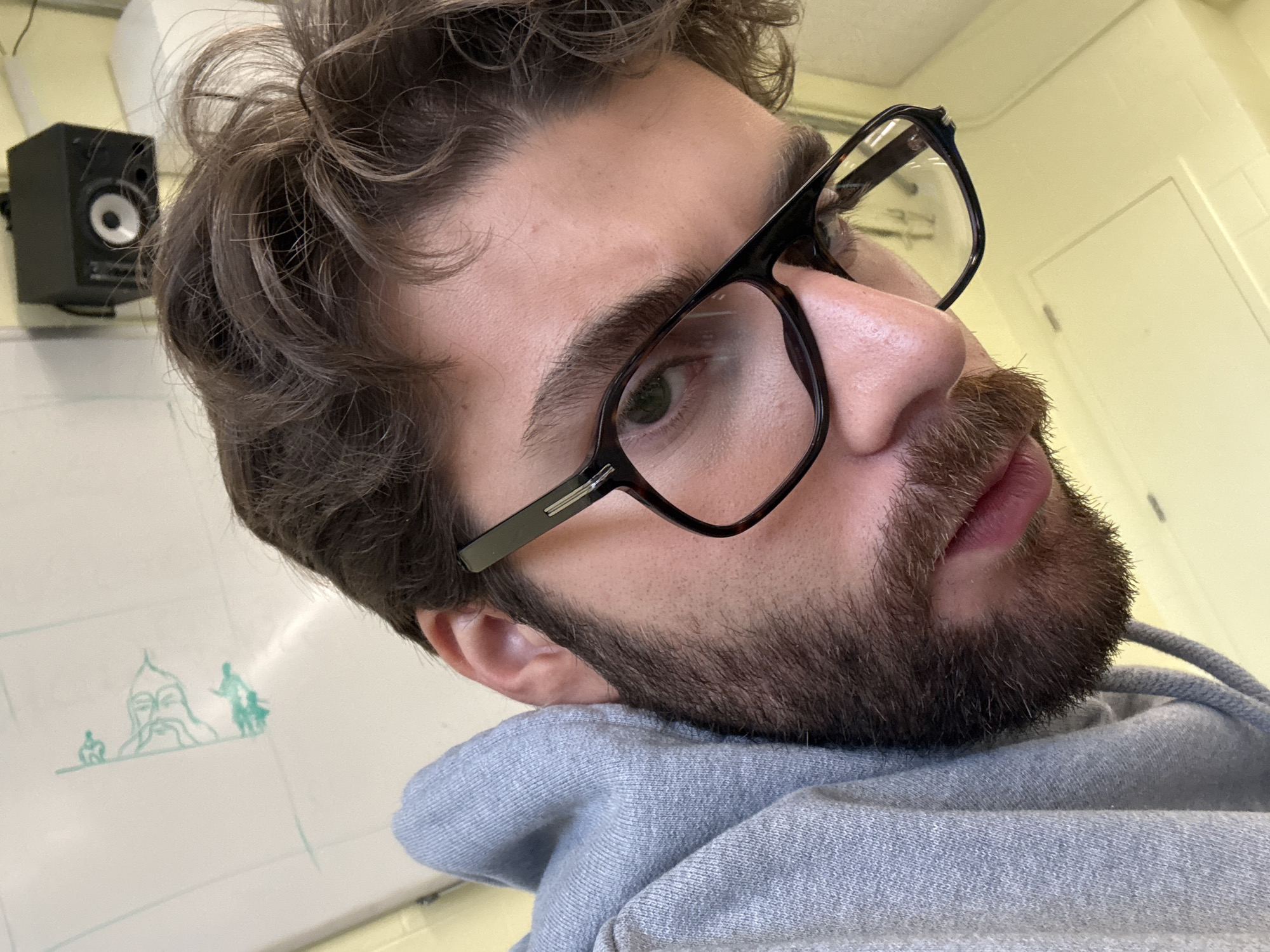 A close-up photograph depicts a person with fair skin, a beard, and brown hair, wearing black-rimmed glasses and a grey hooded sweatshirt. The person is positioned at an angle, with their head tilted towards the left. The focus is primarily on their face and upper body. In the background, there is a white board with a drawing of figures, and a small black speaker mounted on the wall. The location is identified as Montreal, Canada; however, the image does not offer specific details to further identify the scene's location within the city.