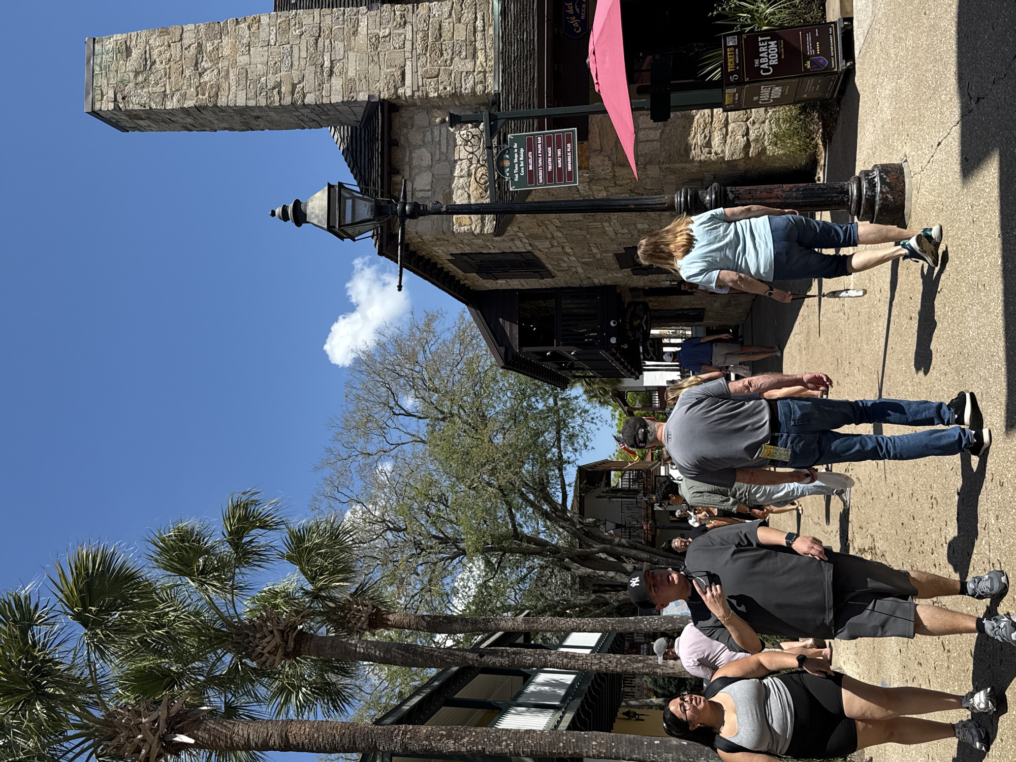 The image depicts a sunny street scene in what appears to be the historic district of Saint Augustine, Florida, United States. Several pedestrians are walking along the paved sidewalk. A woman with blonde hair wears a blue shirt and cropped denim pants and walks near an advertisement sign for "The Cabaret Room." Further along the sidewalk, a man in a gray t-shirt and jeans walks. Closer to the camera, a man wearing a black shirt, shorts, and a baseball cap appears to be taking a photo with his phone. Next to him, a woman wears a gray tank top and black shorts. Palm trees and other trees line the street, with building facades visible in the background. Signs for "Café del Hidalgo" and "The Cabaret Room" are visible. A building with stone construction and a pointed roof is also shown.