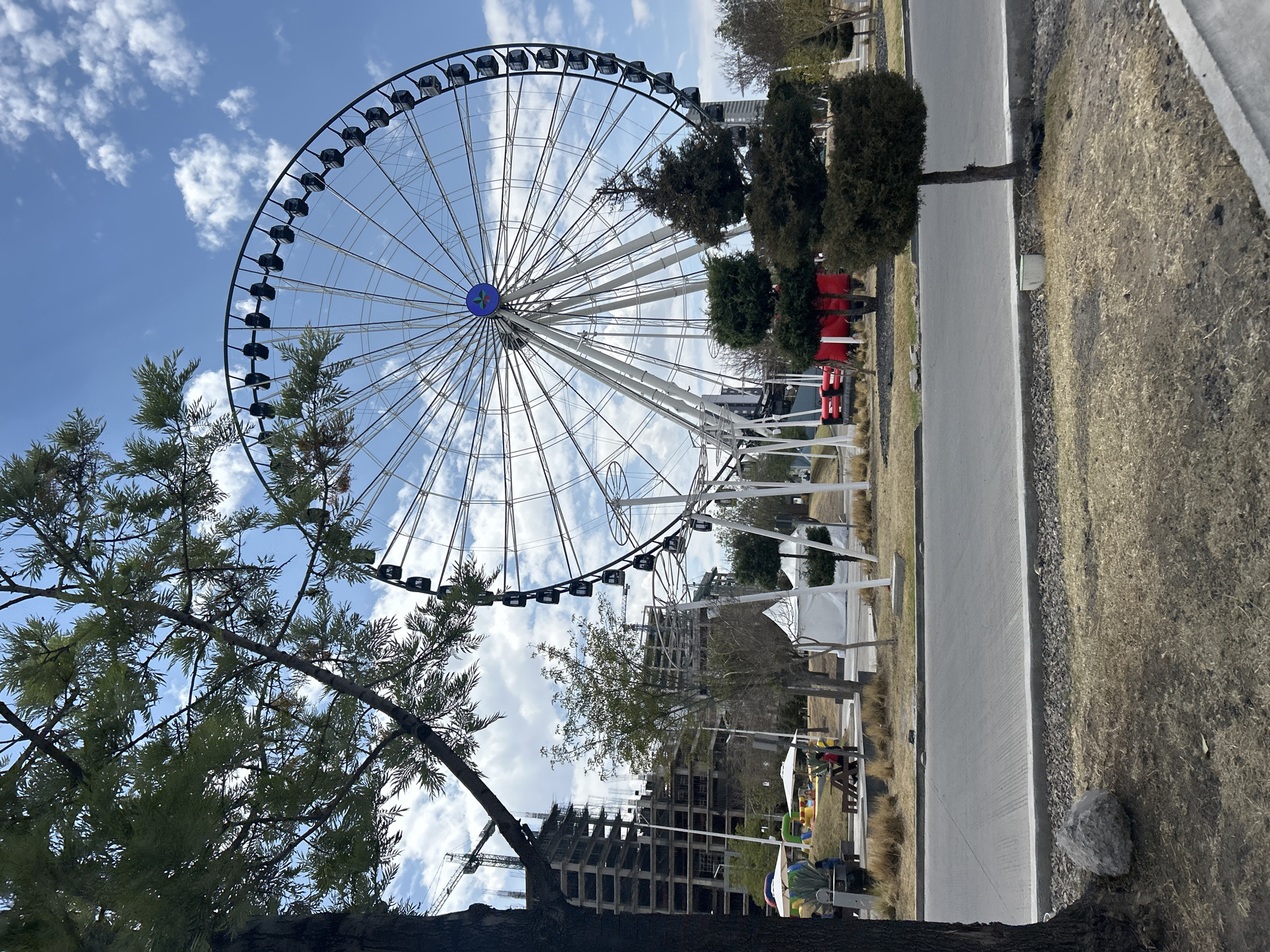 The scene is in San Bernardino Tlaxcalancingo, Mexico. A large Ferris wheel dominates the frame, its structure partially obscured by a tree in the foreground. The sky is partly cloudy, and sunlight filters through the clouds. Next to the road there are a lot of trees and one building is under construction.