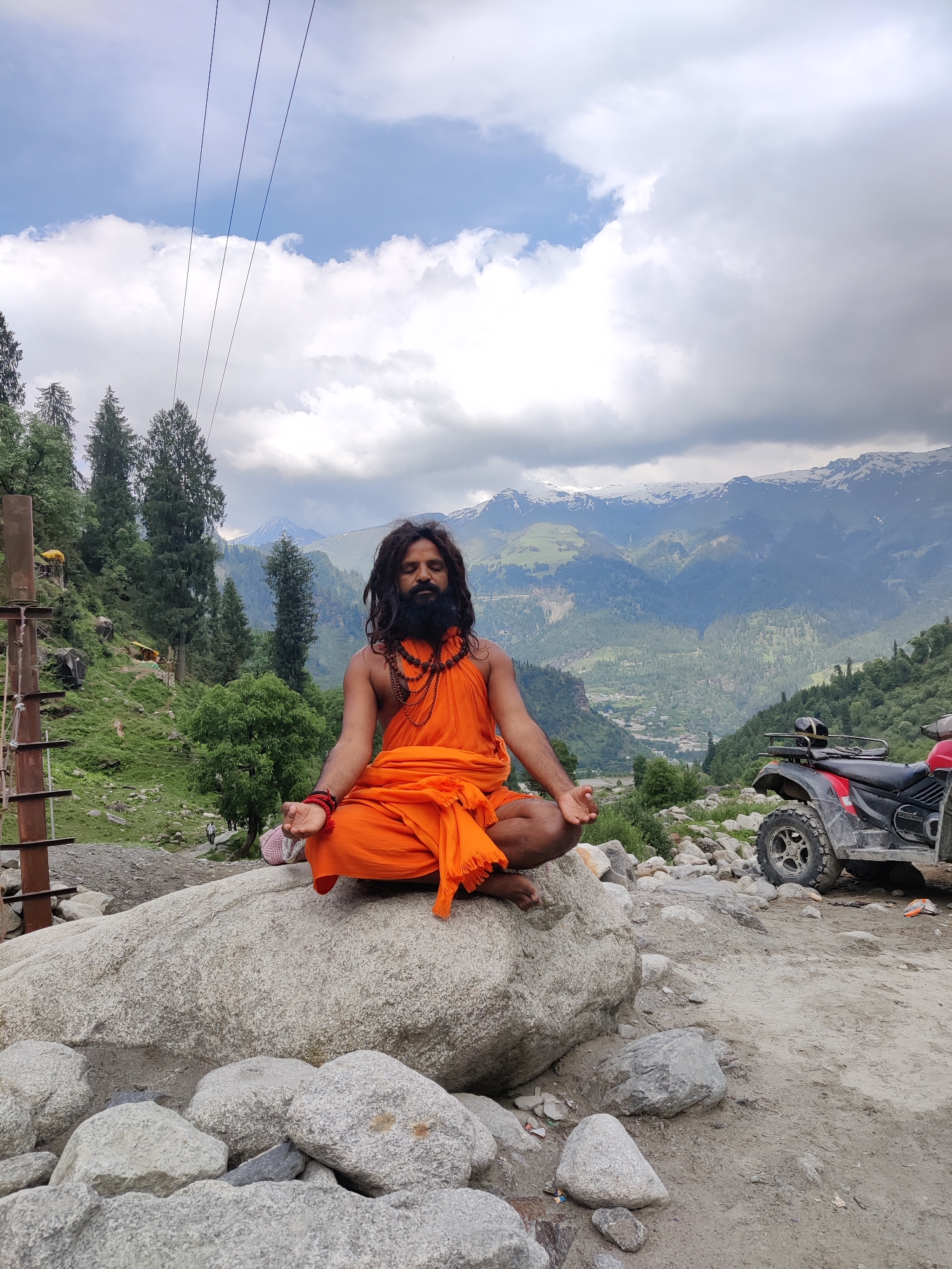 A man in orange robes with a long beard and dark, matted hair sits in a meditative pose on a large boulder. He has prayer beads around his neck and wrists. His eyes are closed, and his hands are resting on his knees with thumbs touching. Behind him, a lush green valley stretches out, dotted with trees and a small village nestled below. Snow-capped mountains rise in the distance, under a cloudy sky. To the right of the frame, a red and black ATV is parked on a dirt path. The scene appears to be in a mountainous, rural area, likely during the daytime, with the weather suggesting it might be cloudy or overcast. The man's posture and expression suggest a state of peace and contemplation. There is a metal pole with some yellow objects attached to it on the left side of the image, and a few small pieces of trash are visible on the ground in the foreground. The location is identified as Majhach, India.