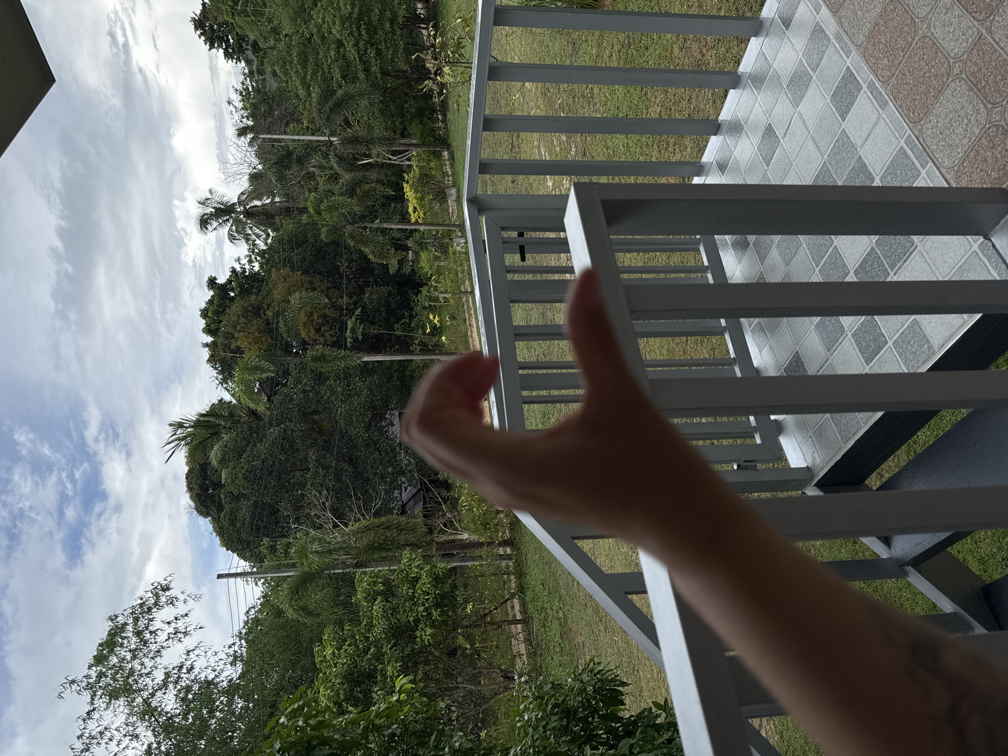 The image depicts a view from a porch or balcony, possibly in Baan Don Sai, Thailand, although this cannot be definitively confirmed from the image alone. The railing of the porch is light gray and constructed of vertical slats. The flooring consists of light gray tiles with a diamond pattern, alongside earth-toned tiles. Outside the railing, a lush green landscape is visible, comprising various trees, including palm trees. The sky is overcast with cloudy conditions. A human hand is prominently positioned in the foreground, obscuring part of the view. Power lines can be seen running through the treeline.