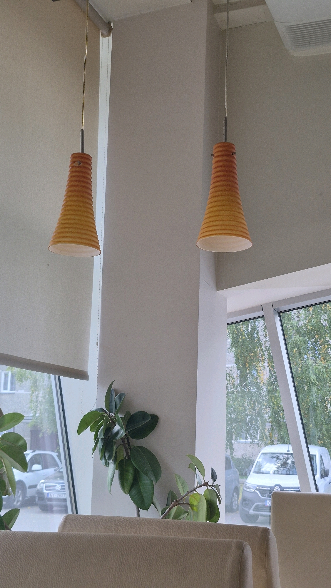 The image depicts the interior of a room with two orange conical pendant lights hanging from the ceiling. To the left, a roller blind is partially lowered, revealing a window with a view of parked cars and buildings. A rubber plant with large green leaves is positioned to the right of the blind, near the window. In the foreground, the tops of two beige upholstered chairs are visible. The environment suggests a cafe or indoor seating area. The specific location in Ludza, Latvia is not explicitly indicated by any visual cues within the image.
