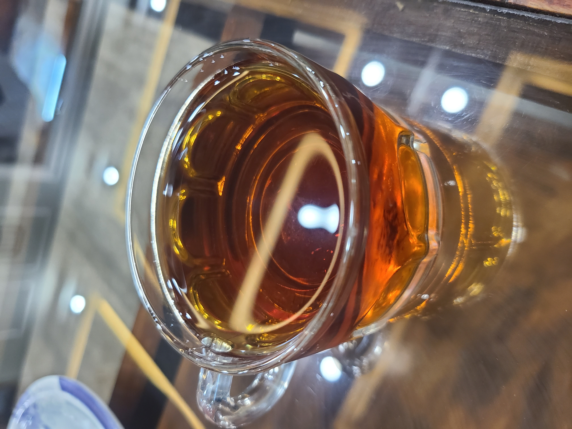 A close-up, high-angle shot shows a clear glass mug filled with a reddish-brown liquid, likely tea. The mug is resting on a glossy, reflective surface, possibly a table, with a visible geometric pattern. The liquid's surface reflects light sources, creating bright highlights. Part of a blue and white object is partially visible in the lower-left corner. The location is Quetta City Tehsil, Pakistan.