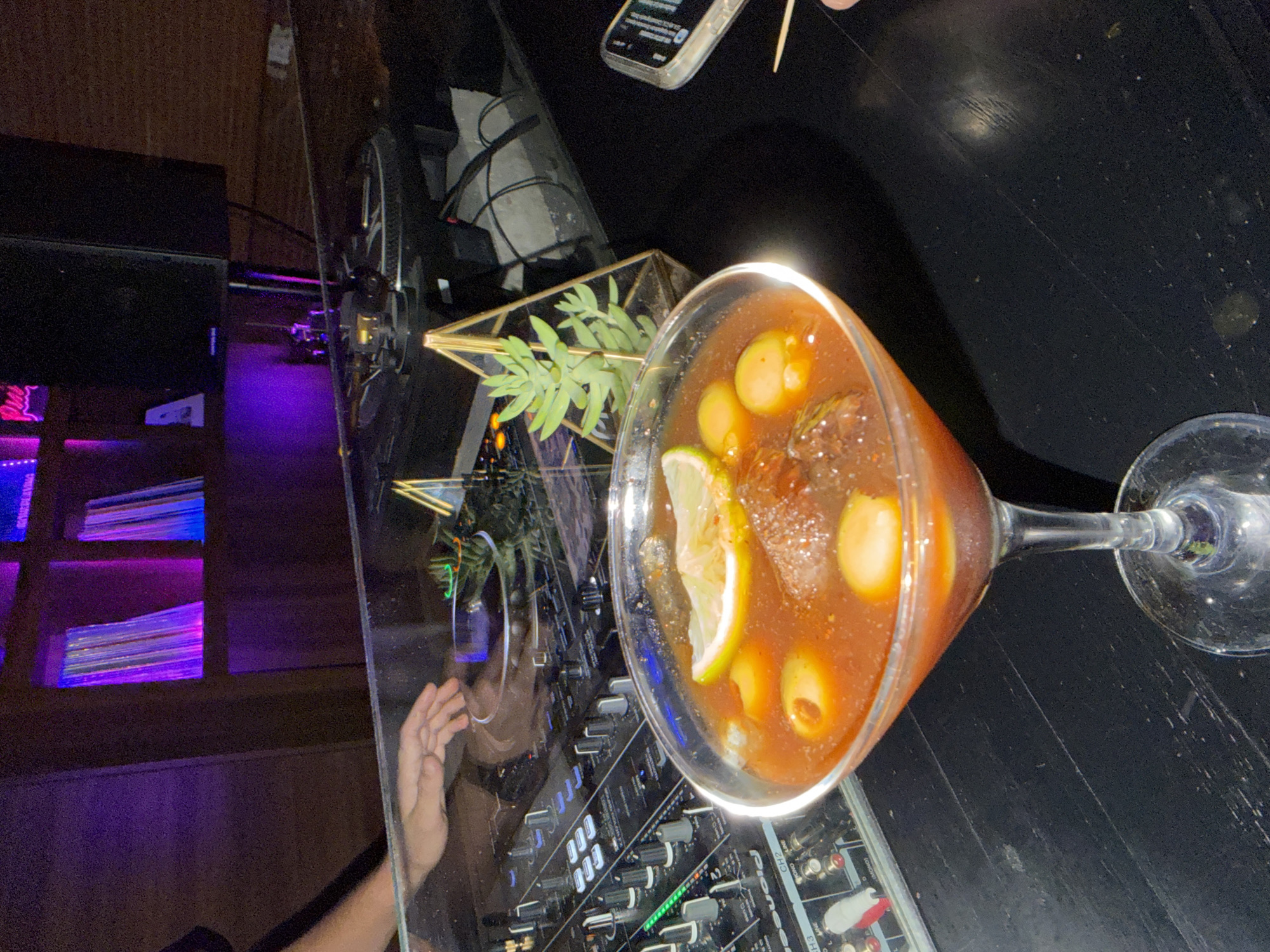 A horizontal, indoor shot shows a cocktail in a martini glass on a glossy black surface next to DJ equipment at a venue in San Andrés Cholula, Mexico. The cocktail is garnished with olives and a lime wedge. A geometric brass and glass terrarium containing green plants sits next to the glass. The left side of the image shows a purple neon-lit display rack and a reflection of a person's hand and arm in the glossy surface. The top of a smartphone is visible in the upper right.