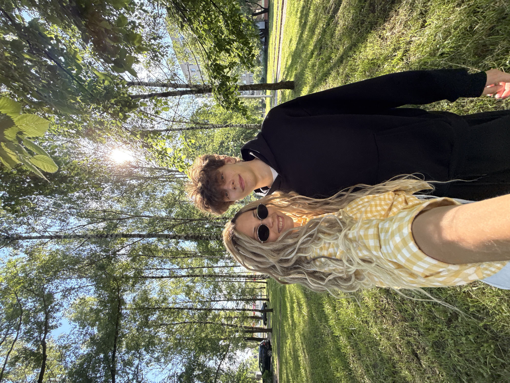 A young woman with blonde dreadlocks and sunglasses smiles at the camera, holding a phone to take a selfie. She is wearing a yellow and white checkered shirt. Beside her, a young man with curly brown hair smiles slightly. He is wearing a black hoodie. They are standing in a park with tall trees and green grass. The sun is shining through the trees, creating lens flare. In the background, cars are parked along a street, and a building is visible. The scene appears to be in an urban park on a sunny day.