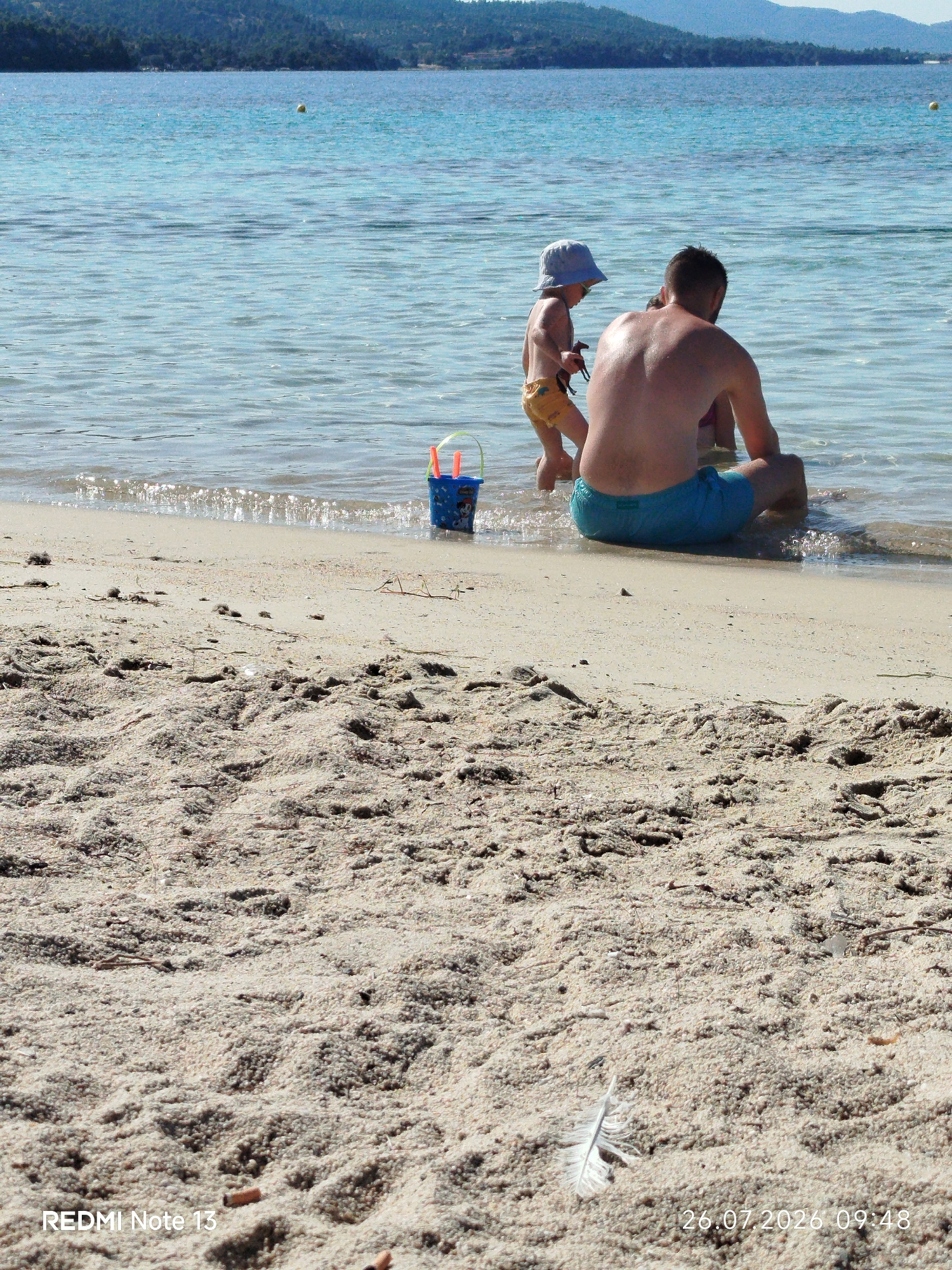 A father and his young child are playing in the shallow, clear blue water at a sandy beach in Sithonia Municipal Unit, Greece. The father, wearing blue swim trunks, is seated in the water facing away from the camera, with his back glistening from the water. The child, wearing yellow shorts and a light blue hat, stands to the left of the father, facing the water and holding a toy. A blue plastic bucket with orange toys sits on the wet sand at the water's edge. The beach is covered in light-colored sand with some scattered debris and a single white feather in the foreground. In the background, the calm sea stretches towards a tree-covered coastline and distant hills under a clear sky. The bottom right corner displays a date and time stamp: "26.07.2026 09:48".
