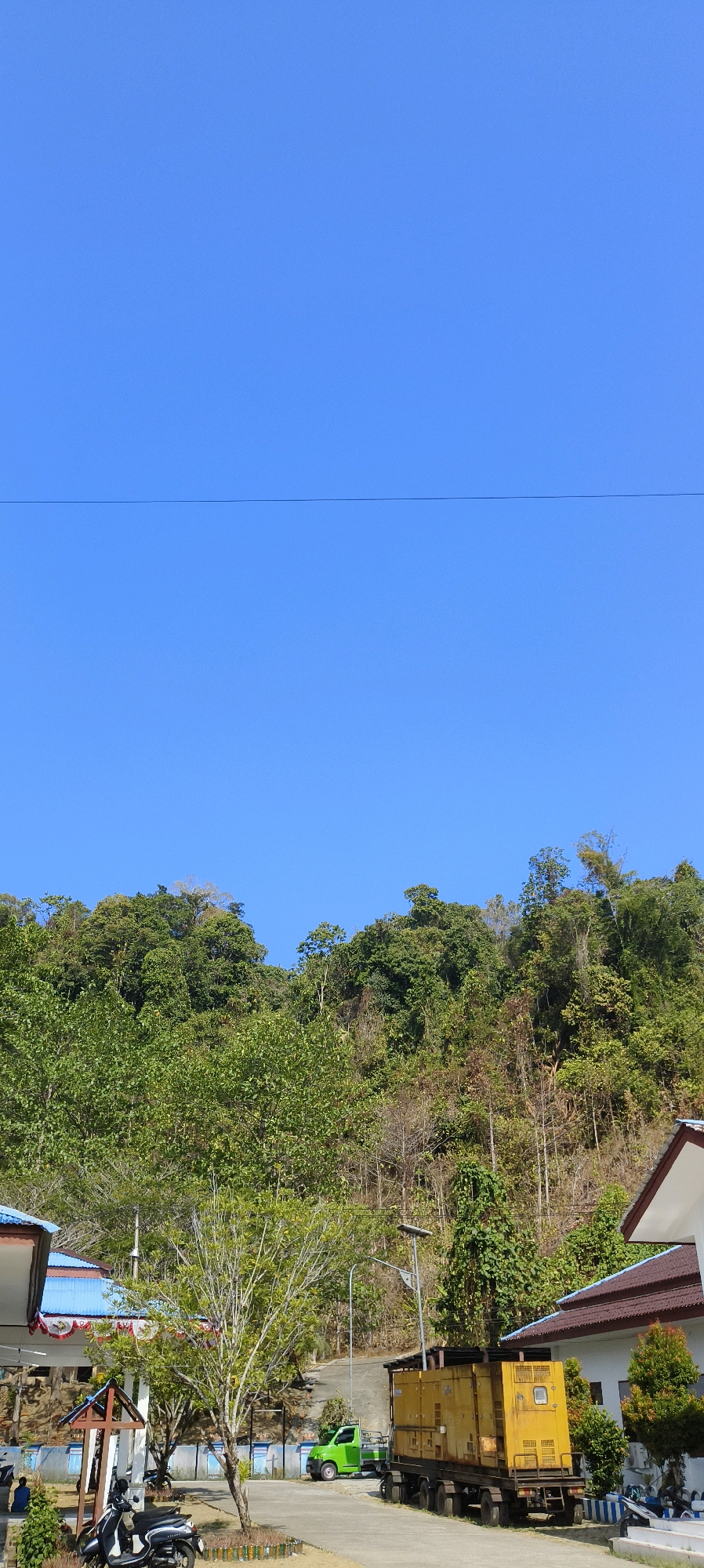 The image captures a bright, clear day in a semi-rural area of Waisai, Indonesia, characterized by lush greenery and a vibrant blue sky. In the foreground, a portion of a white building with a brown roof is visible on the right, suggesting a residential or administrative structure. Adjacent to it, a large, yellow generator unit is parked on a paved surface, hitched to a trailer with multiple wheels. To the left of the generator, a small, lime green pickup truck is parked.
A slender tree with sparse green leaves stands near the center of the frame, its branches reaching upwards. Several motorcycles are parked in the shaded area near a low-lying building with a distinctive blue roof adorned with white trim. This building appears to be a pavilion or an open-air structure. Further into the background, a hillside covered in dense green foliage rises steeply, with a clear demarcation between the vegetated area and the bright sky. A thin, dark power line stretches horizontally across the upper section of the sky. The overall impression is one of a sunlit, relatively quiet location, likely on the outskirts of Waisai, where infrastructure blends with the natural landscape. There are no visible people actively engaged in any significant actions, though a figure is seated in the shade of the building on the far left.