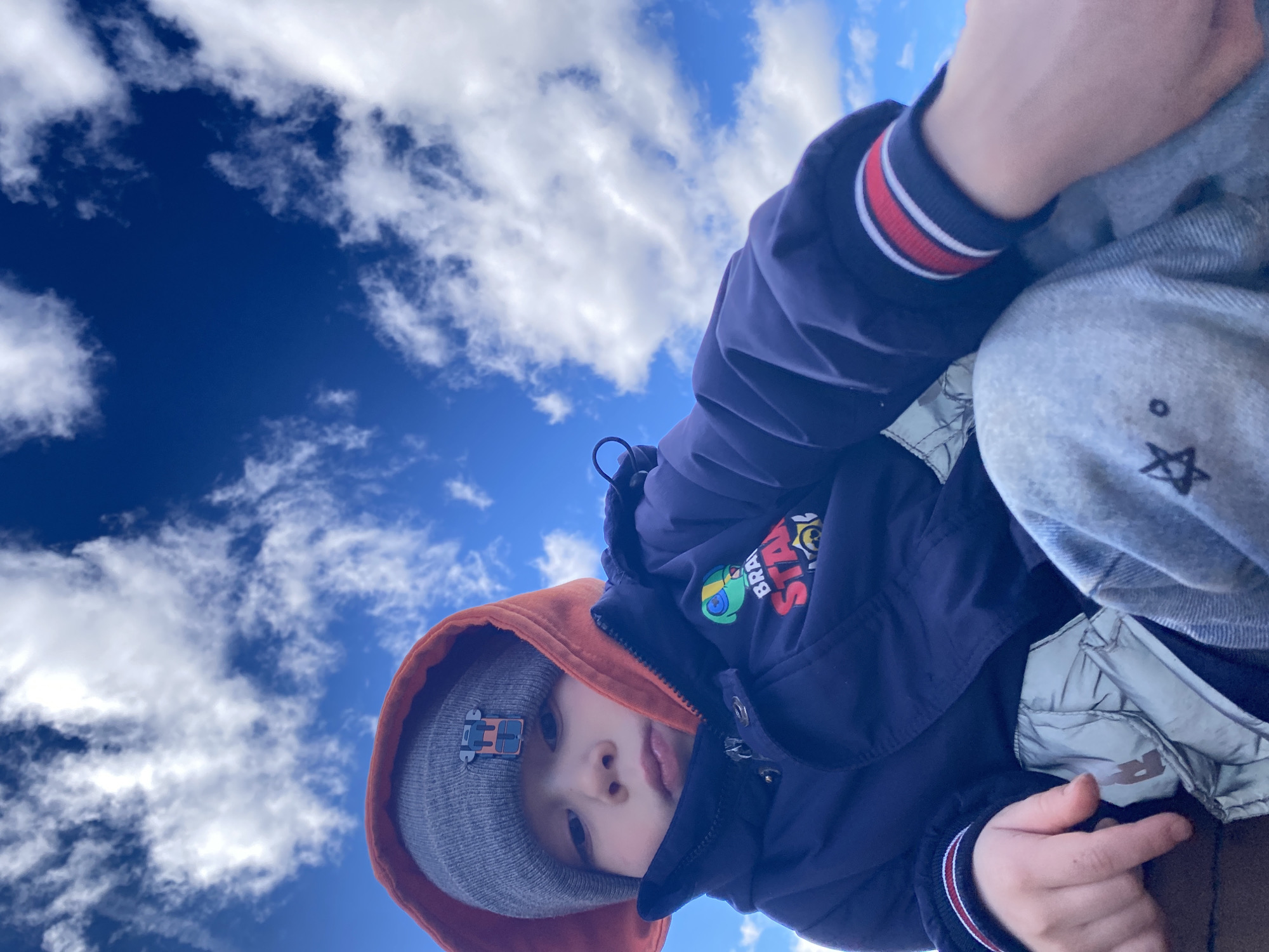 A low-angle shot captures a child from below, seemingly crouching or leaning over the camera. The sky is visible in the background. The child wears a grey beanie with a small toy attached, an orange hoodie, and a navy blue jacket with red and white stripes on the sleeve. The jacket also has a patch with the words "STAR BRA" visible. The child is also wearing grey pants with a white star design. The background shows a clear blue sky with scattered white clouds. There is no indication of Ольховка, Russia.