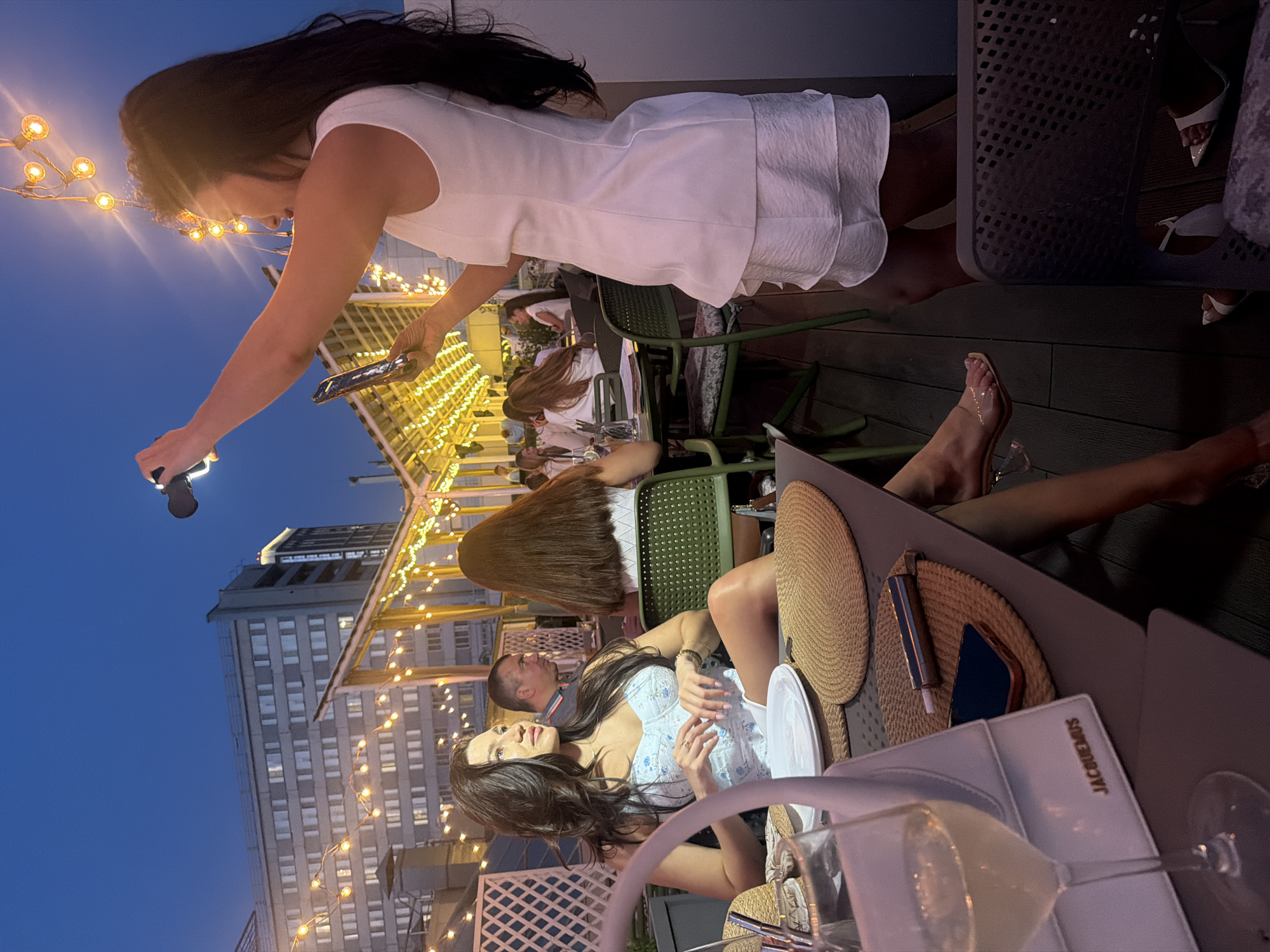 The image captures a group of people socializing on a rooftop patio at dusk in Novosibirsk, Russia. The scene is illuminated by string lights and the ambient glow of the city. In the foreground, a woman with long dark hair, wearing a white dress, is holding a phone and a black object that appears to be a microphone or remote. She is looking down, possibly recording or directing something. Nearby, another woman, seated and wearing a light blue corset-style top, is engaged in conversation with a man. She has long dark hair and is looking to her left.
Several other individuals are visible in the background, seated at tables. One woman with long brown hair is partially obscured. The setting suggests a restaurant or bar with outdoor seating, featuring metal chairs and tables. The architecture of the buildings in the background indicates an urban environment. A tall, modern building with many windows is prominent. The time of day is evident from the deep blue sky and the artificial lighting, suggesting it is evening. The overall mood seems relaxed and social, with people enjoying each other's company. The visible text on a white object in the foreground reads "JACQUEMUS."