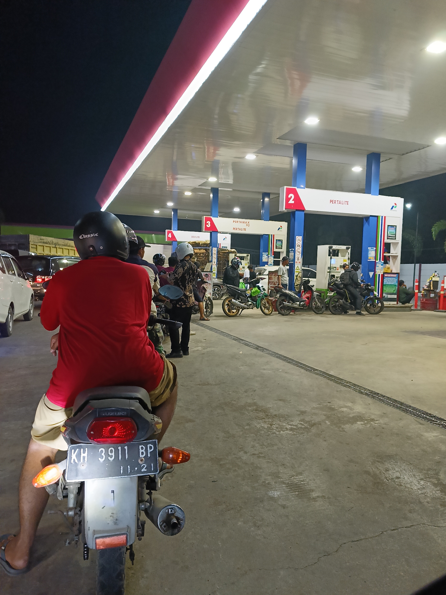 This is a vertical, eye-level shot of a busy gas station in Palangka Raya, Indonesia, at night. The foreground is dominated by the back of a person sitting on a motorcycle, wearing a red shirt and khaki shorts, with a black helmet. The motorcycle's license plate reads "KH 3911 BP" and the inspection sticker shows "11 . 21".
The gas station canopy extends across the midground, illuminated by bright white lights. Two gas pumps are visible under the canopy, with signage indicating "2 PERTALITE". Further back, under another section of the canopy, a pump is marked with "3 PERTAMAX 92" and "PERTALITE". Several other motorcycles and a white car are parked or waiting at the pumps. People are seen interacting with the pumps and on their motorcycles. The ground is concrete, with a metal grate running along the lower right portion of the image. The background is dark, with some palm trees and distant buildings visible. The scene suggests a typical evening at a gas station with multiple customers.