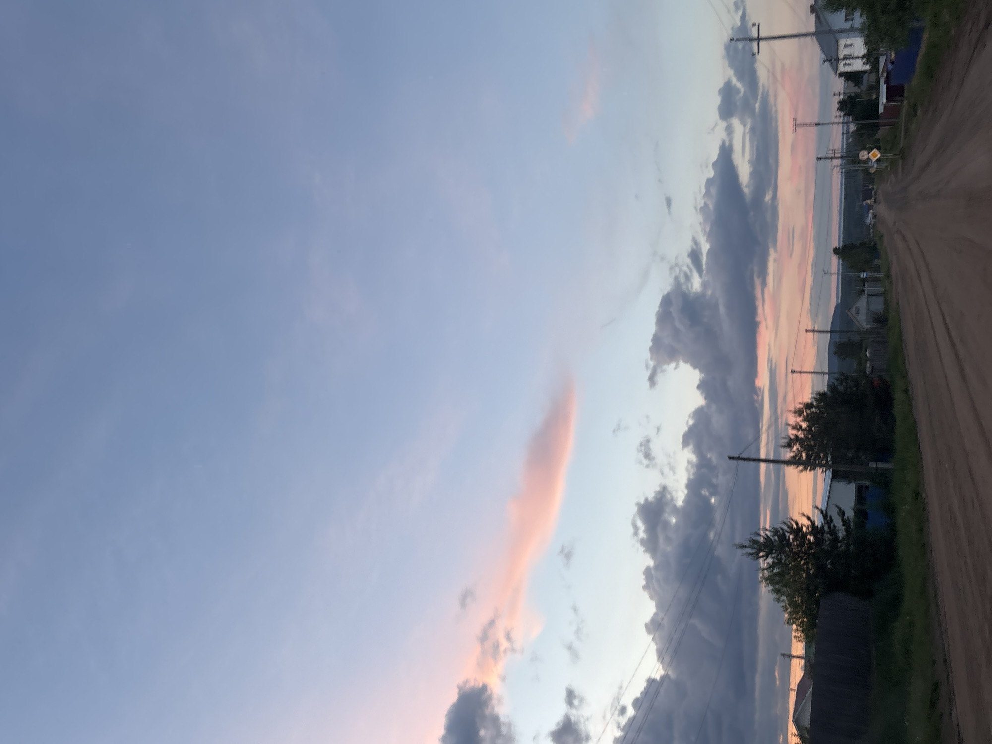 The image shows a dirt road in Ольховка, Russia, flanked by houses and vegetation. Utility poles and power lines run along the road. The sky transitions from blue to pink and gray, with various cloud formations. There are no people visible in the image.