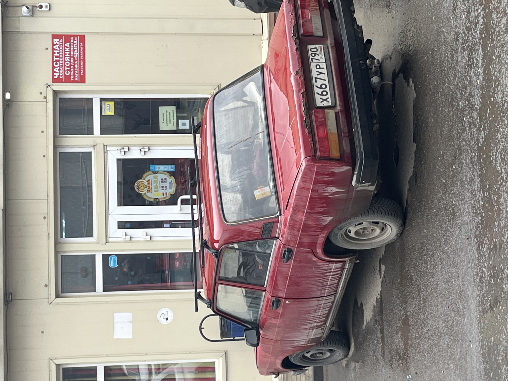 The image captures a red Lada sedan parked sideways on a wet surface. The car's license plate is "X667YP 790," indicating it is registered in Russia. The car's body shows signs of rust and wear.
The car is parked near a building, which appears to be a store. A sign on the building reads "ЧАСТНАЯ СОБСТВЕННОСТЬ" (Private Property), "СТОЯНКА" (Parking), "только длЯ КЛИЕНТОВ МАГАЗИНА «ЦЫПА»" (Only for customers of the "Tsypa" store), and "РАБОТАЕТ ЭВАКУАТОР" (Evacuator works). A sticker reading "HEBA" is also on the door frame. A store hours sign reads "9.00-20.00"
Based on the Cyrillic text on the signage, the image is likely taken in a Russian-speaking region. The license plate indicates that the car is registered in Russia. Assuming the store name "ЦЫПА" is a local store and the license plate is authentic, and that "HEBA" is a local store, this is likely the city of Klin, Russia.
