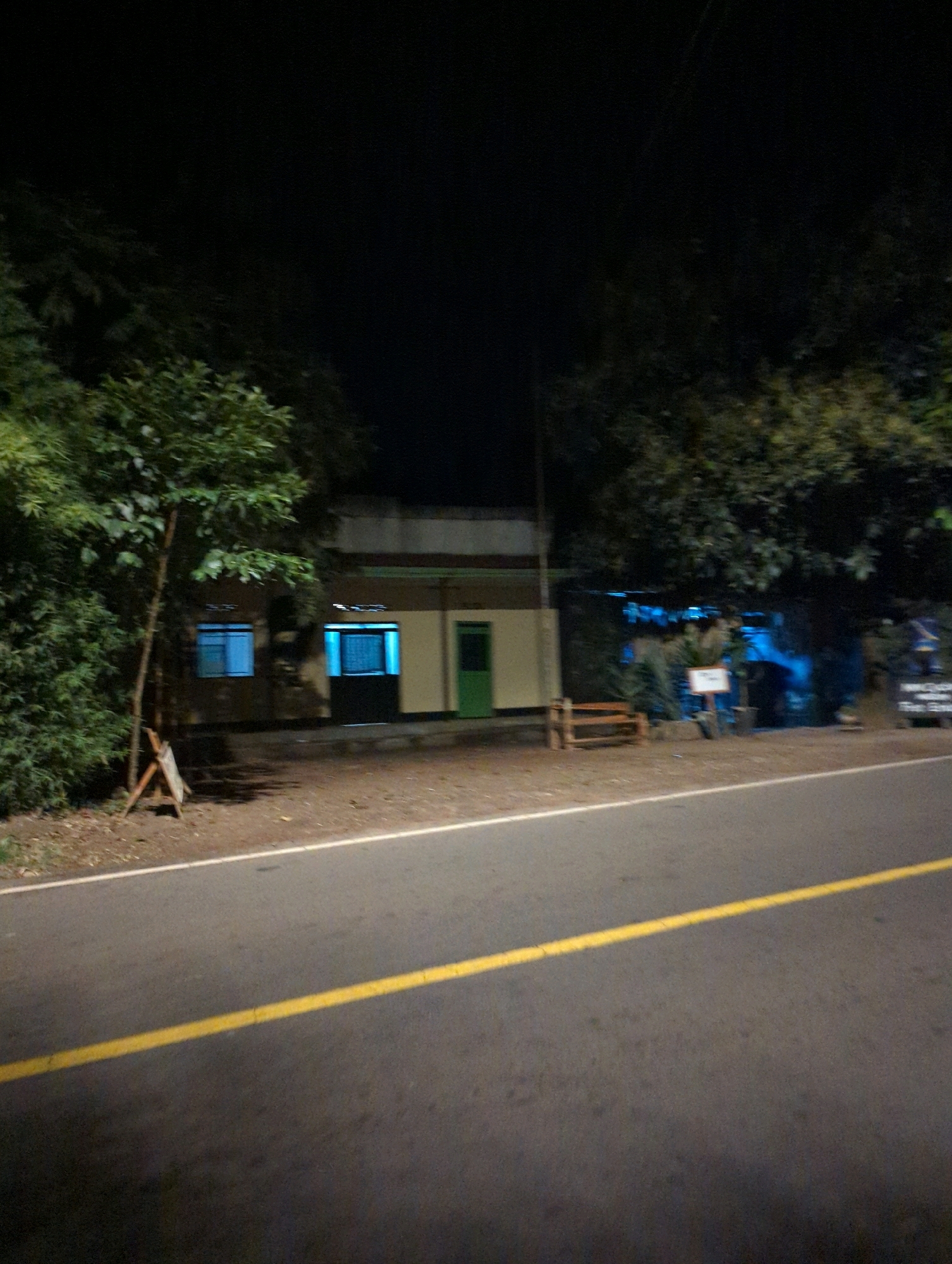 A night scene captures a roadside building in Musanze District, Rwanda. The building has a light-colored facade with two windows emitting a bright blue light, and a green door on the right. In front of the building, a wooden bench and a small wooden easel are visible. Trees and foliage surround the structure, with the dark night sky overhead. The foreground shows a paved road with a white line and a single yellow line marking the lane. The scene is dimly lit by artificial lights from the building and possibly surrounding areas.
