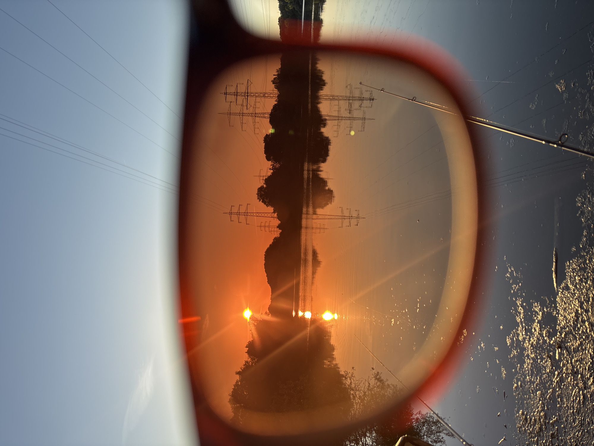 The image shows a sunset viewed through sunglasses. The sunglasses have red frames and tinted lenses that give the scene an orange hue. In the reflection on the water, there are two fishing rods. In the background, there are trees, a body of water, and power lines with tall pylons. The scene appears to be a body of water in or near Ludwigshafen am Rhein, Germany, given the presence of power lines and pylons often found in industrial or semi-industrial areas. The time of day is sunset, indicated by the low sun and the orange and pink colors in the sky and its reflection on the water.