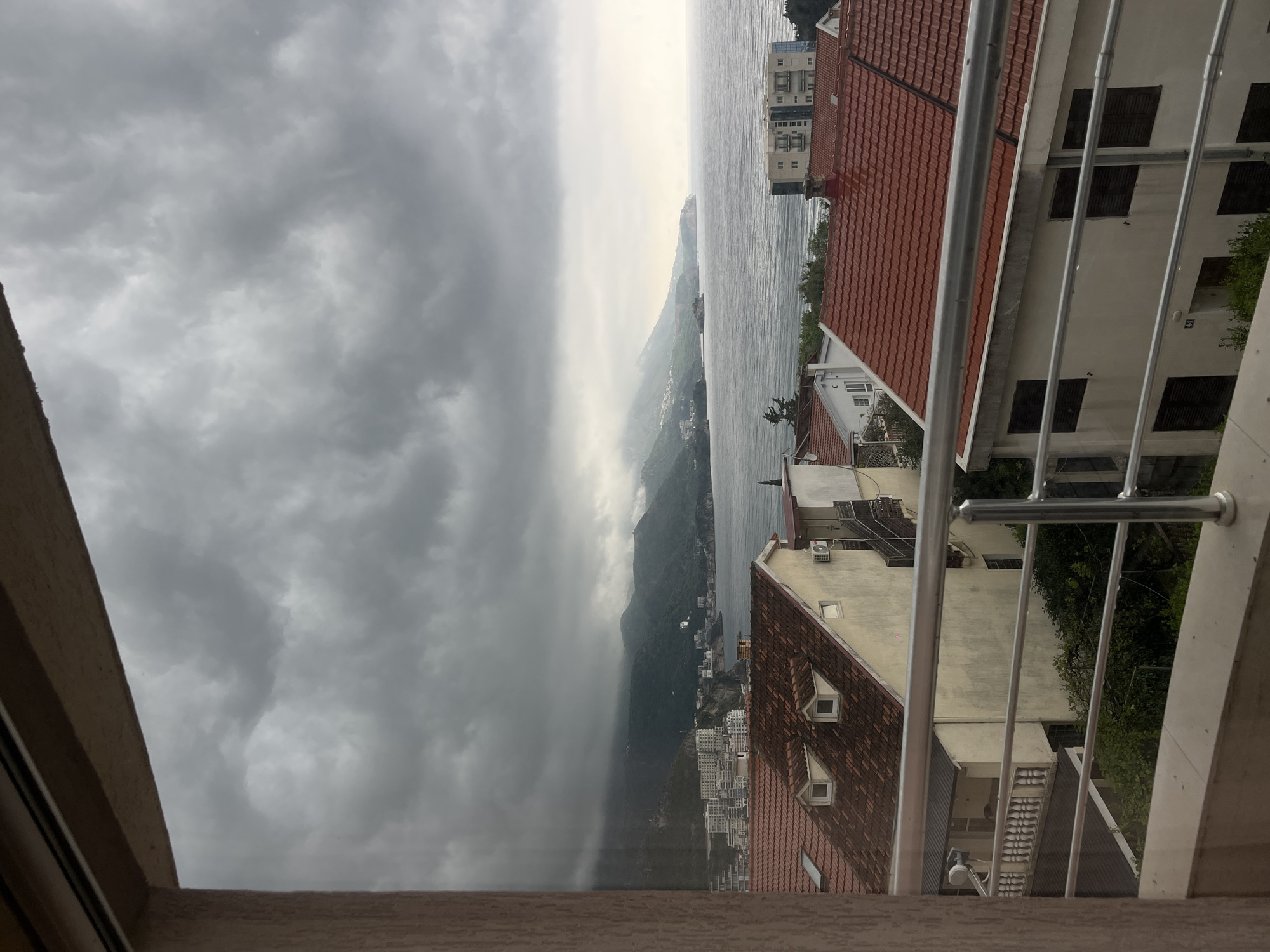 The image captures an overcast coastal landscape, likely from a balcony or elevated window in Boreti, Montenegro. The scene is dominated by a vast, grey, and uniformly cloudy sky that blends into a hazy horizon over a large body of water, possibly the Adriatic Sea.
In the immediate foreground on the right, a shiny, silver metal railing of a balcony is prominently featured. Below and behind this railing, parts of several buildings are visible, characterized by their reddish-brown tiled roofs. Some of these structures are light-colored with visible windows, and a few feature small, distinct dormer windows on their roofs.
Beyond the foreground buildings, the calm, grey expanse of water stretches into the midground. The distant coastline is marked by rolling hills or mountains that rise from the sea. These slopes are dotted with numerous smaller buildings, appearing as white clusters against the dark green of the terrain. The entire distant landscape is shrouded in a soft haze, making the more remote features indistinct and merging them with the overcast sky.
The weather is clearly cloudy and appears damp, contributing to the muted color palette and subdued lighting of the entire scene. There are no people visible, and no specific activity is taking place, presenting a tranquil yet somber view of the coastal environment. No visible text is discernible in the image.