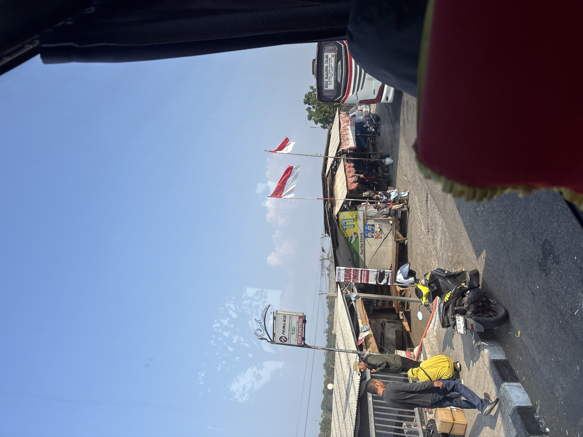 The image depicts a street scene in Cikampek, Indonesia, likely at a bus stop or rest area. Two Indonesian flags fly on poles in front of a row of small shops and stalls. A white and red bus with "BEKASI - MAJALENGKA - CINJUR" printed on its rear is parked on the right side of the road. In the foreground, a person wearing a dark long-sleeved shirt and jeans walks on a sidewalk next to some stacked boxes. A yellow and black motorcycle is parked on the street. Several other individuals are visible near the shops, some sitting and others standing. The sky is clear blue with some scattered clouds. The overall environment suggests a busy but casual commercial area.