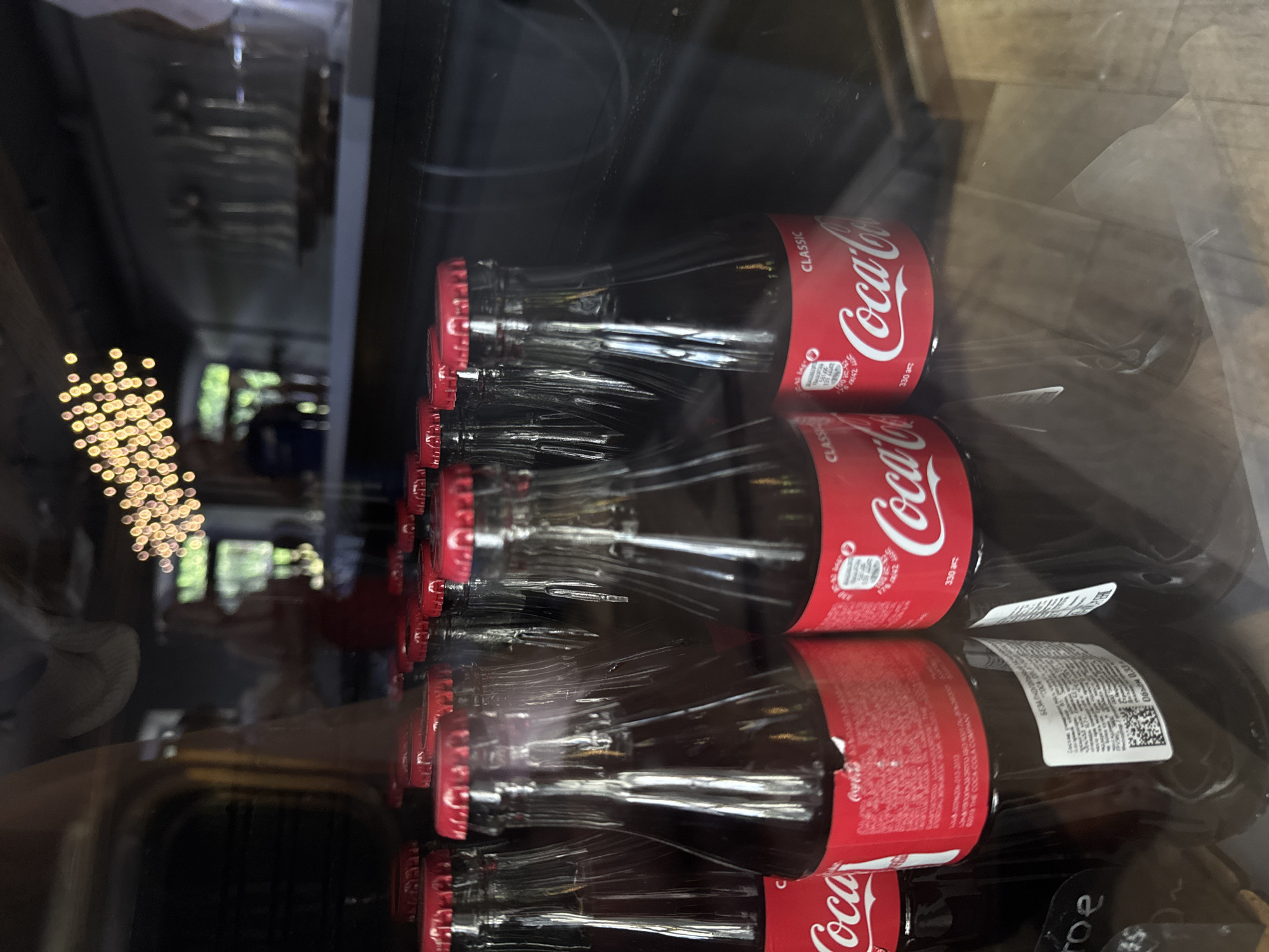 The image captures multiple glass bottles of Coca-Cola Classic, arranged in rows, likely within a refrigerator or display case. The bottles are oriented horizontally due to the image's rotation. Each bottle features the iconic red label with "Coca-Cola" prominently displayed in white script, along with "CLASSIC" and "330 ml" (or "330 мл" in Cyrillic).
The glass surface of the display reflects the surrounding environment, creating a layered visual effect. In the background, out-of-focus golden lights, possibly string lights or decorative bulbs, create a bokeh effect, suggesting an indoor setting, potentially in the evening or night. Blurred shapes of an interior room are also visible in the reflections, along with a hint of greenery, possibly from a window or an indoor plant.
No people are visible, and no specific activity is taking place beyond the static display of the beverages. The reflections, especially the golden lights, convey a warm or cozy ambiance, despite the subject being a commercial product.
Visible text includes:
- "Coca-Cola"
- "CLASSIC"
- "330 ml" (or "330 мл")
- "БЕЗАЛКОГОЛЬ" (Non-alcoholic in Cyrillic/Russian)
- "Объём 0.33" (Volume 0.33, referring to liters, also in Cyrillic/Russian)
- "21.3.2010" (a date, likely manufacture or expiry)
- Partially visible text such as "ABOUT THE COCA-COLA COMPANY".
The presence of Cyrillic text like "БЕЗАЛКОГОЛЬ" and "Объём 0.33" aligns with the location context of Izhevsk, Russia.