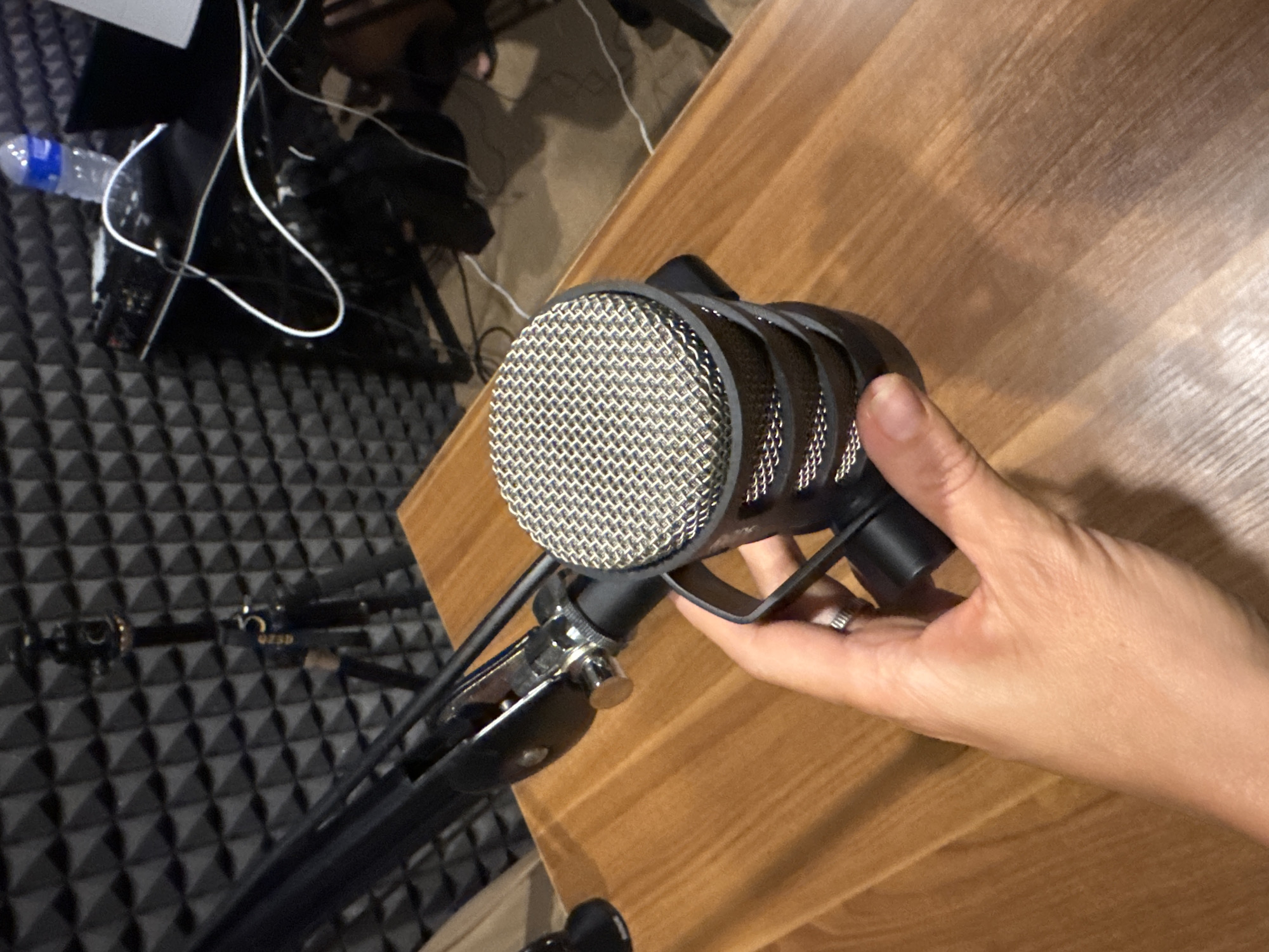 A person's hand is visible holding a microphone mounted on a stand. The microphone is black with a silver-colored grill. A wooden surface is behind the microphone and the hand. The background consists of a black, textured acoustic panel. A water bottle and other objects are visible in the periphery, suggesting an indoor studio or recording environment. The image does not contain enough information to identify the location in Denpasar, Indonesia.