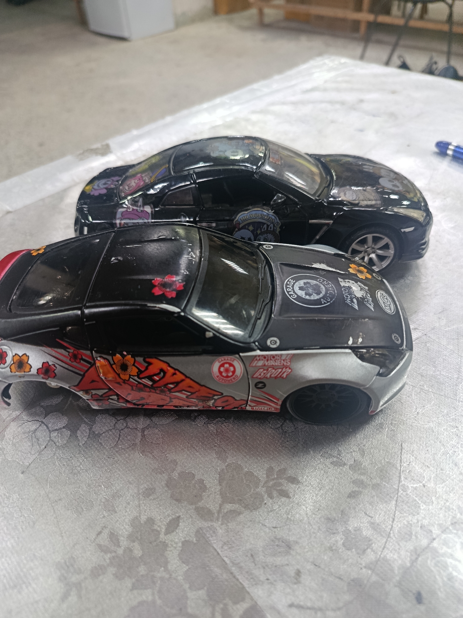 Two toy cars are on a patterned surface. The car in the foreground is black and silver with red and orange decals of flowers and what appear to be tribal designs. The car in the background is black and has various stickers on it. The surface the cars are on is a light-colored, textured fabric with a floral pattern. The background is blurred but appears to be an indoor setting with some furniture. There is no information in the image to suggest a location in Sertolovo, Russia.