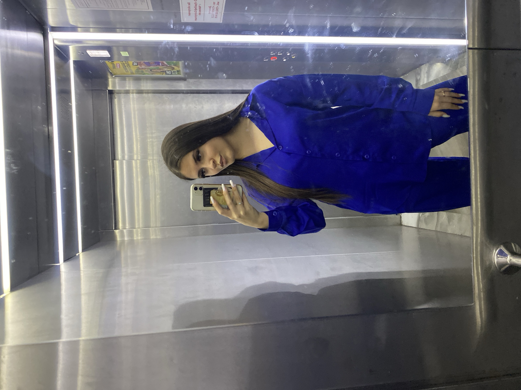 A woman is taking a selfie in an elevator. She is wearing a blue button-down shirt and pants. The elevator has metal walls. There is a sign in Russian on the top wall of the elevator. Based on the signage in Russian, the city is Chaikovsky, Russia.